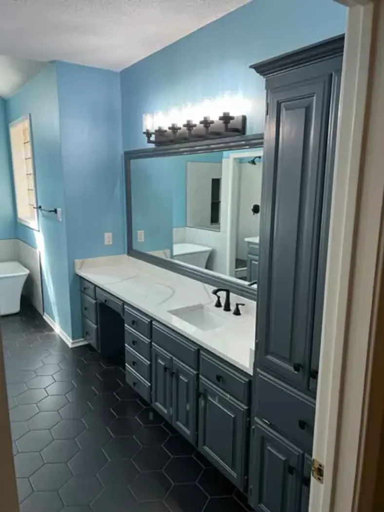 Blue-walled bathroom with dark blue cabinets, white countertops, large mirror, and black hexagon tile floor.