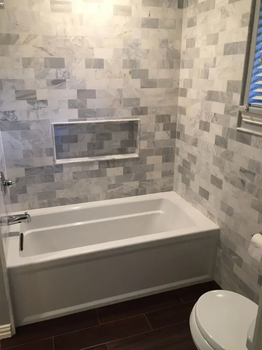Bathroom with white tub, gray and white tiled walls, and wood-look flooring.