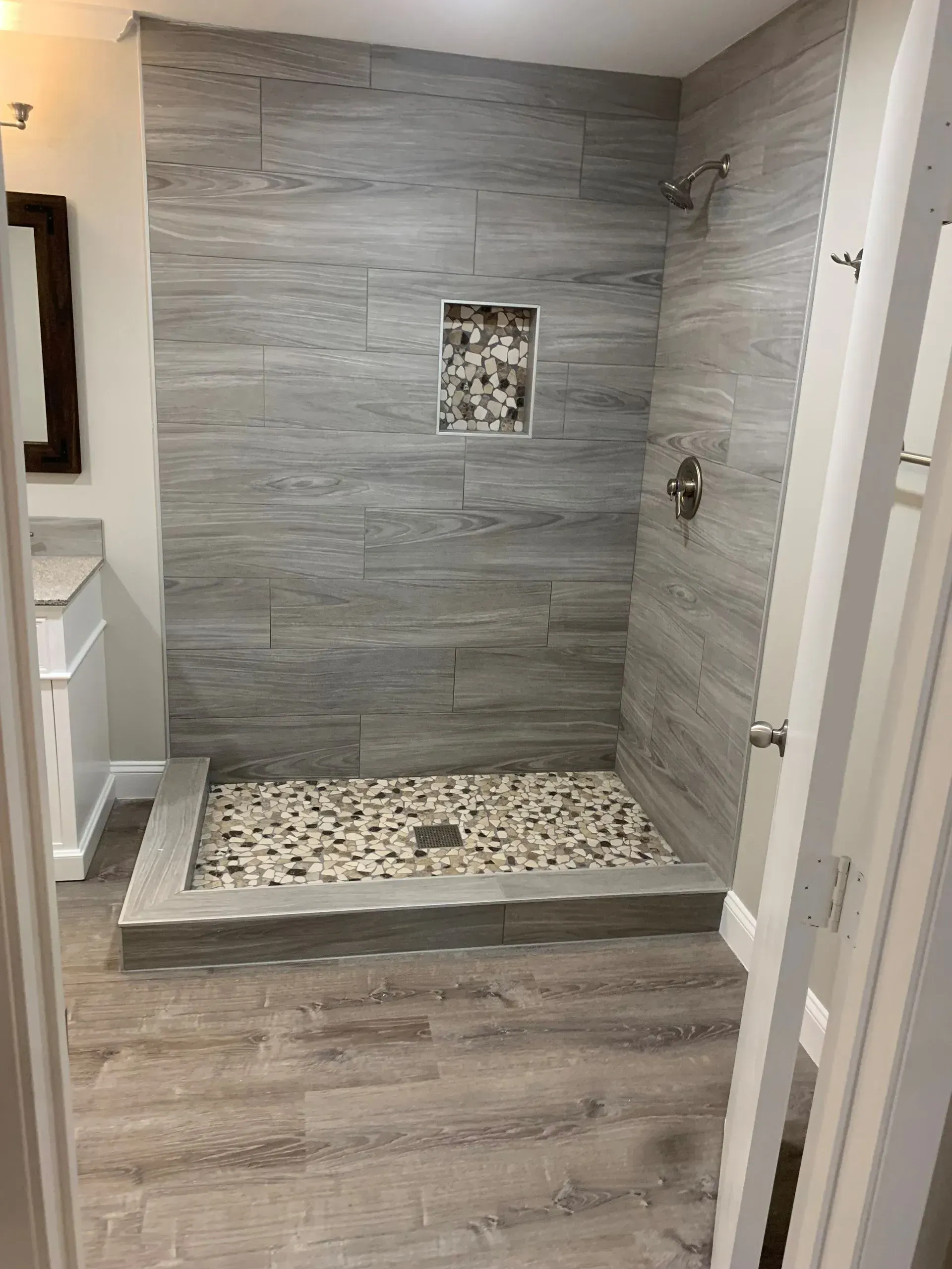Modern bathroom shower with gray tile walls, stone floor, and light wood flooring.