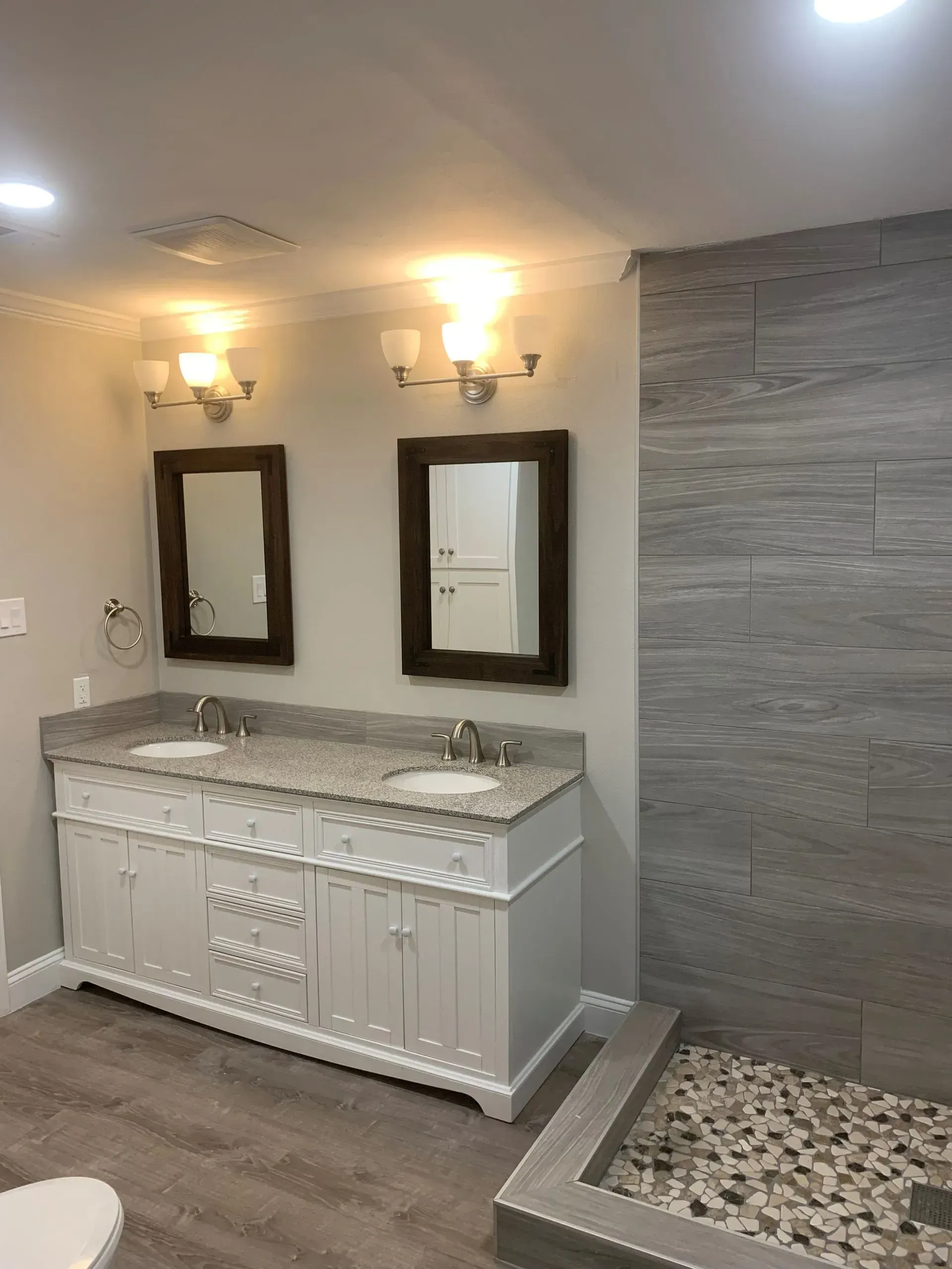 Bathroom with double vanity, two mirrors, and a stone-tiled shower. White cabinets, gray countertops, and light gray walls.