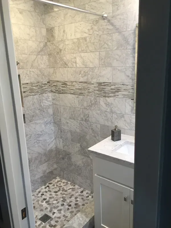 Small bathroom with gray marble tiled shower, pebbled floor, white vanity, and soap dispenser.