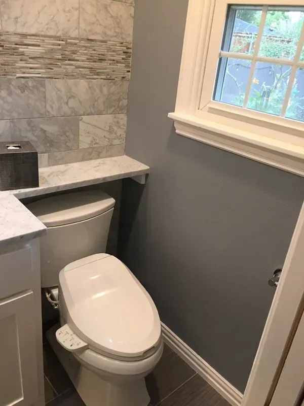 Small bathroom with a toilet, shelf, and window. Gray wall, white trim, and tiled wall.