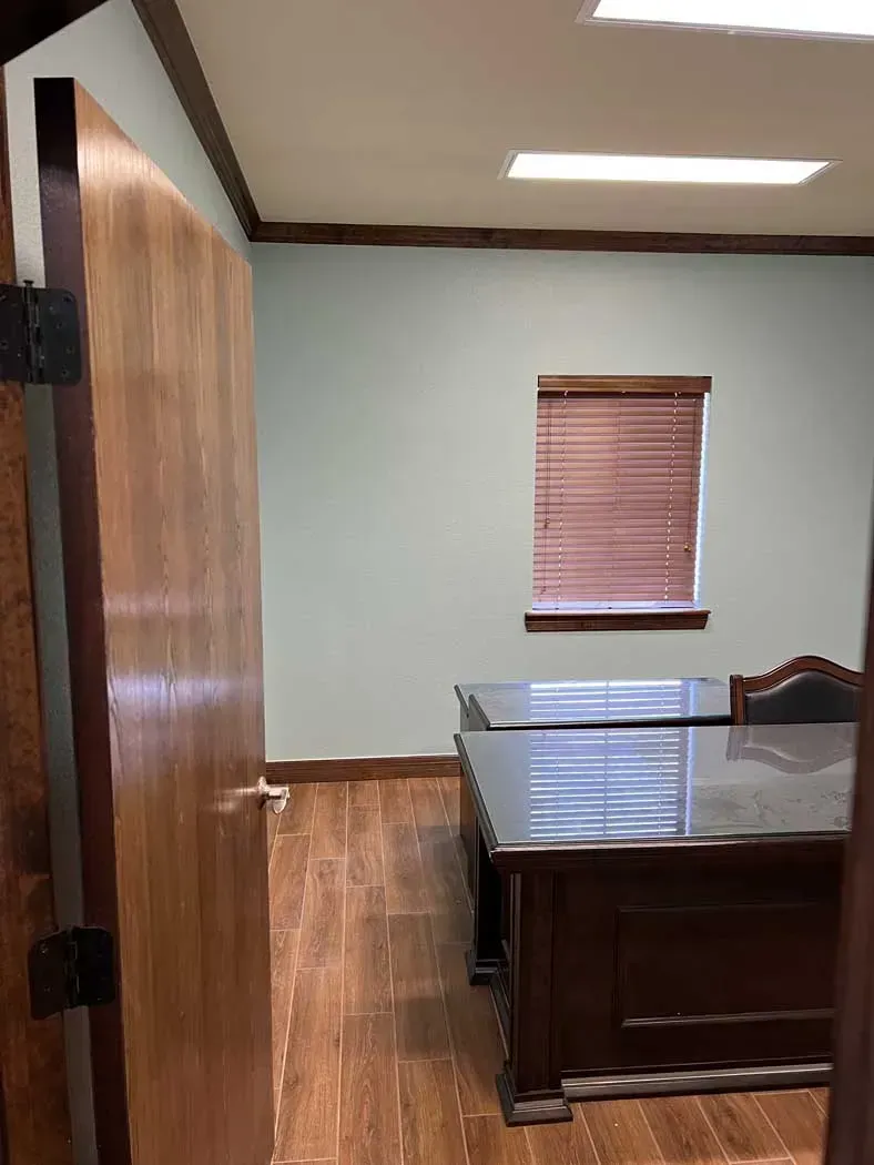 Doorway view of an office with a desk, window, and wooden flooring. Light blue walls and brown trim.