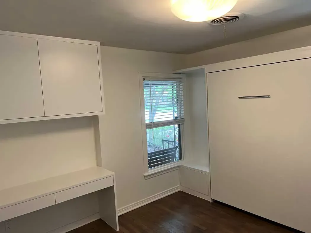 White room with desk, cabinets, window with blinds, and a closed wall bed.