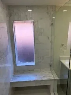 Shower stall with marble bench and window; glass door; light gray and white tiles.