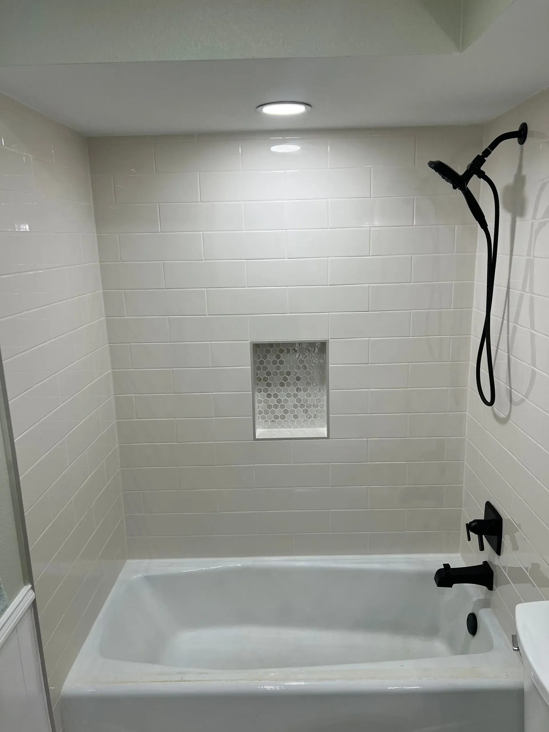 Bathroom with white bathtub, light beige tiled walls, dark fixtures, and recessed shelf.
