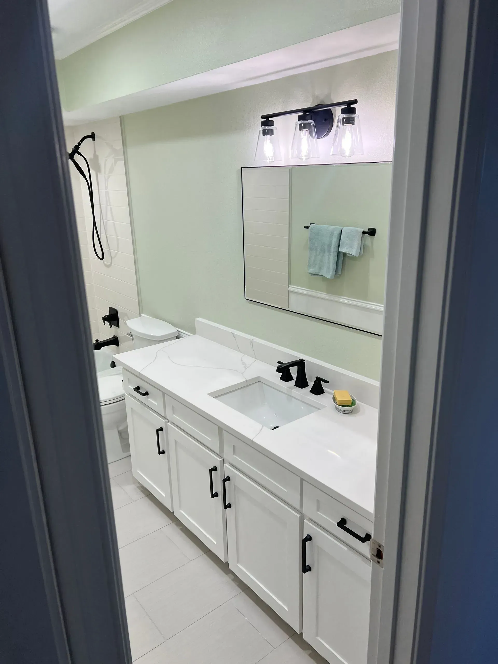 Bathroom with white cabinets, countertop, and fixtures, black hardware, light green walls, and a large mirror.