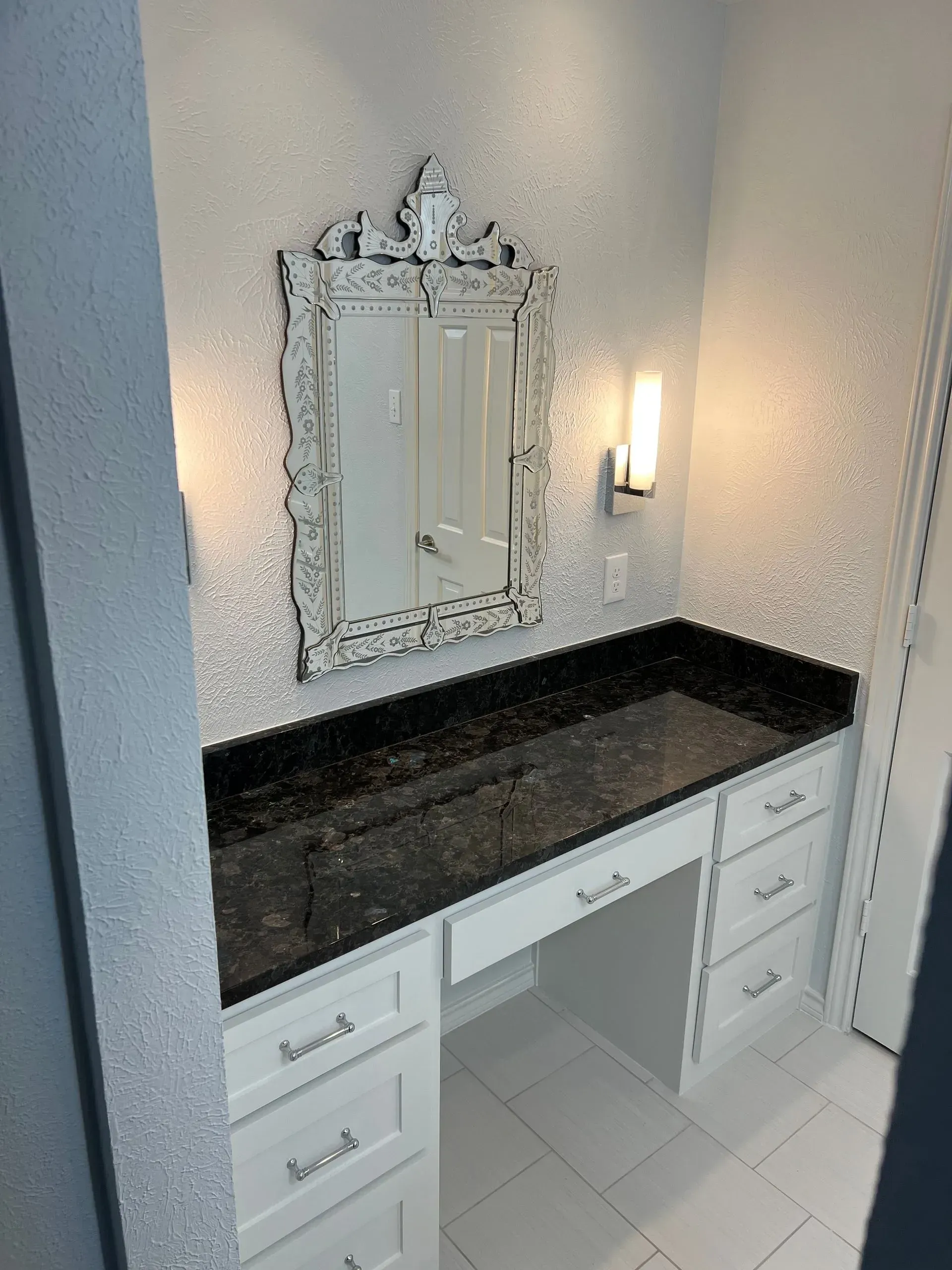 Vanity with black countertop, white cabinets, ornate mirror, and textured wall in a bright space.
