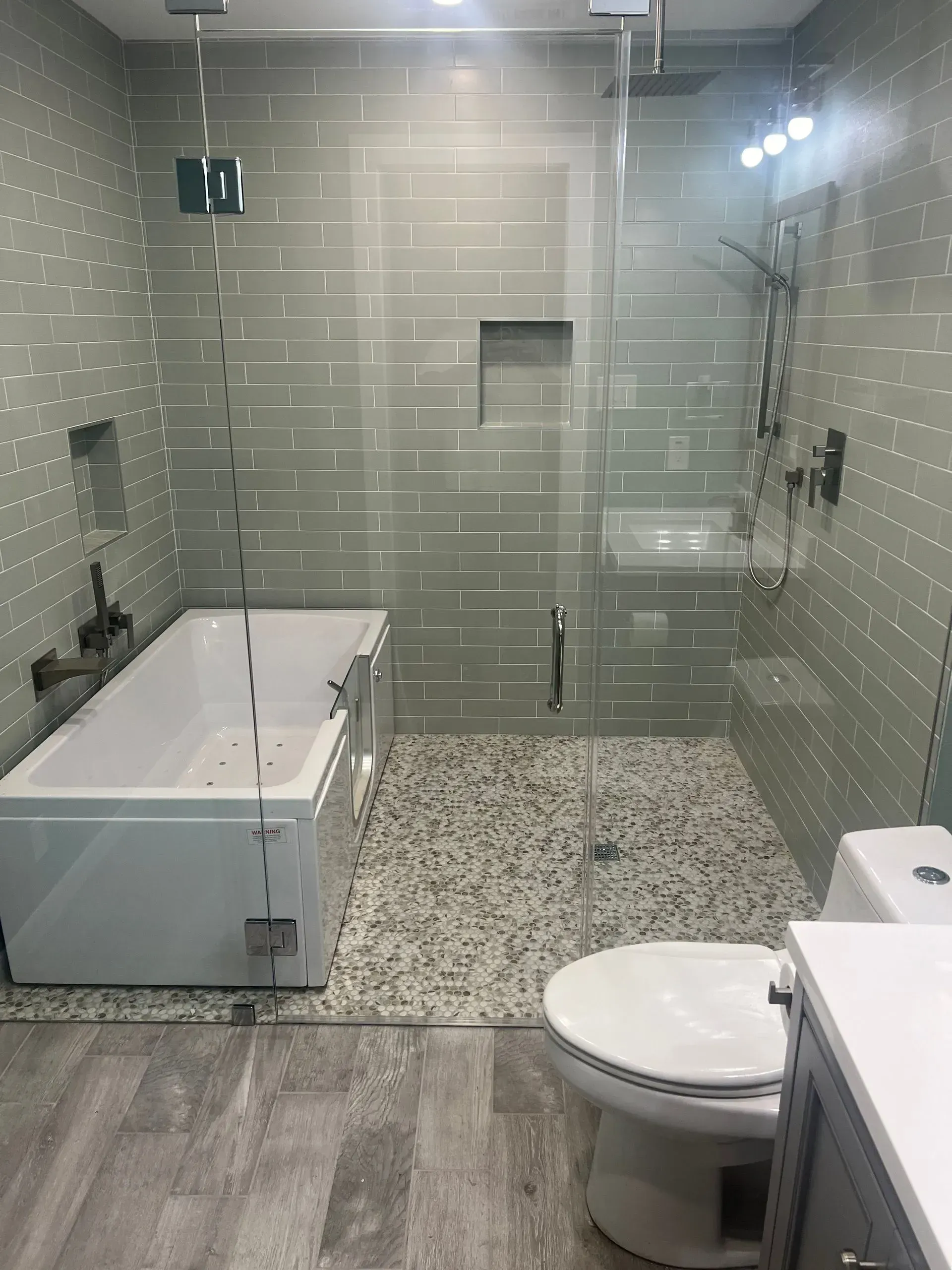 Modern bathroom with glass shower, tub, pebble floor, and white toilet. Gray tile walls and light gray floor.