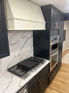 Modern kitchen with dark cabinets, white range hood, and marble backsplash. Stainless steel ovens.