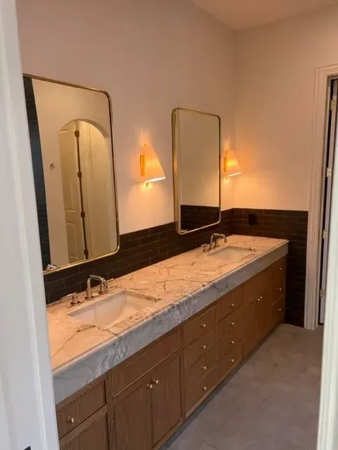 Bathroom with two sinks, two mirrors with sconces, wooden cabinets, and dark tile backsplash.