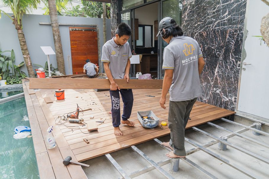 Workers assembling a wooden deck frame on a patio beside a house