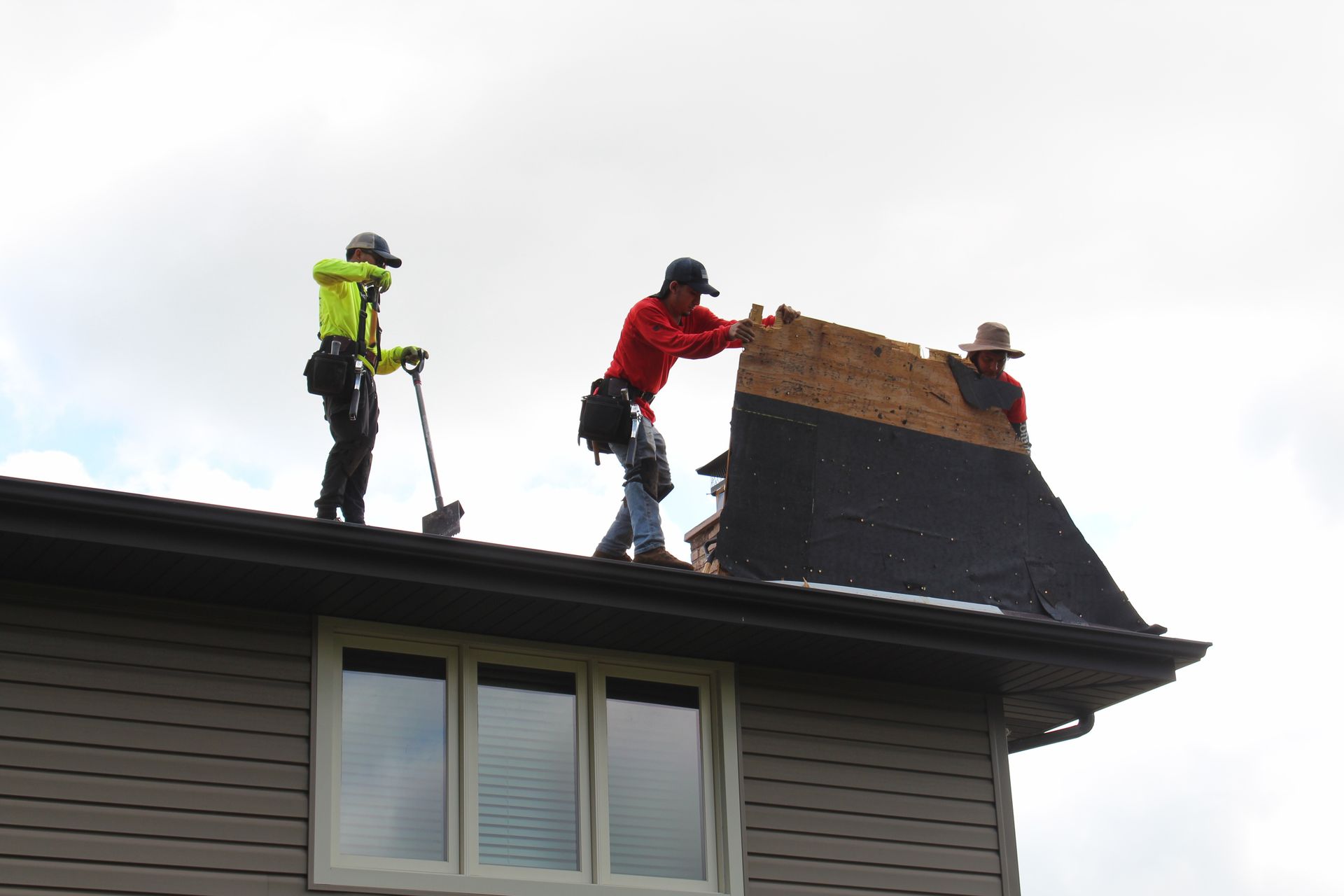 Three roofers on a rooftop removing shingles; cloudy sky.