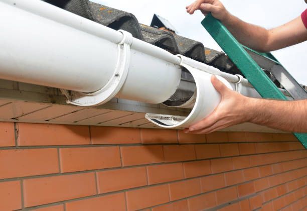 Person installing white gutter, with ladder, on a brick building with dark roof.