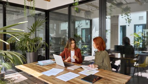 Two women are sitting at a table in an office talking to each other.