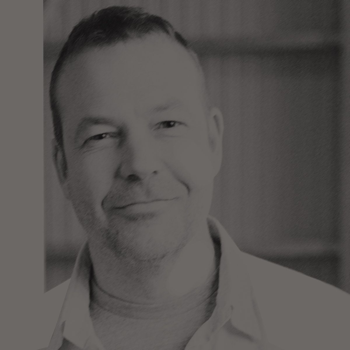 Man with short hair smiles, wearing gray shirt under open white button-down. Wooden beam and corrugated metal background.
