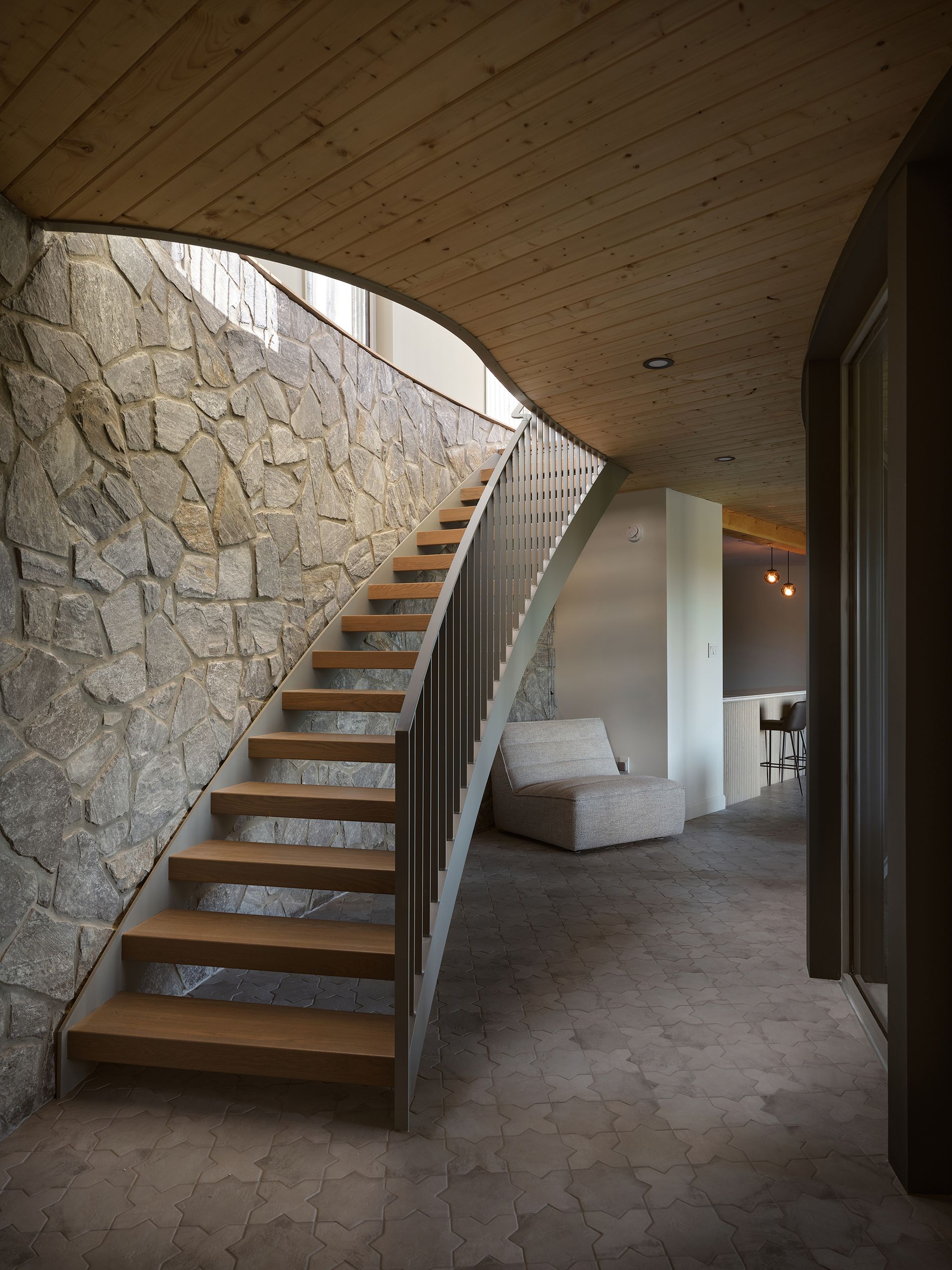 Staircase with wooden steps and metal railing against stone wall, leading upward. Interior view with curved wall, soft seating.