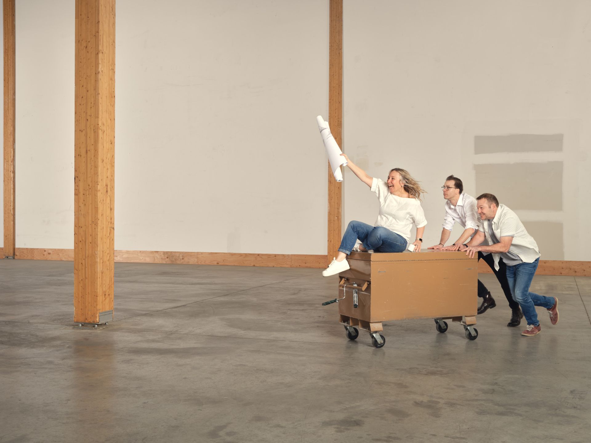 Woman rides cardboard box cart, two people push, inside empty room, holding a paper up.