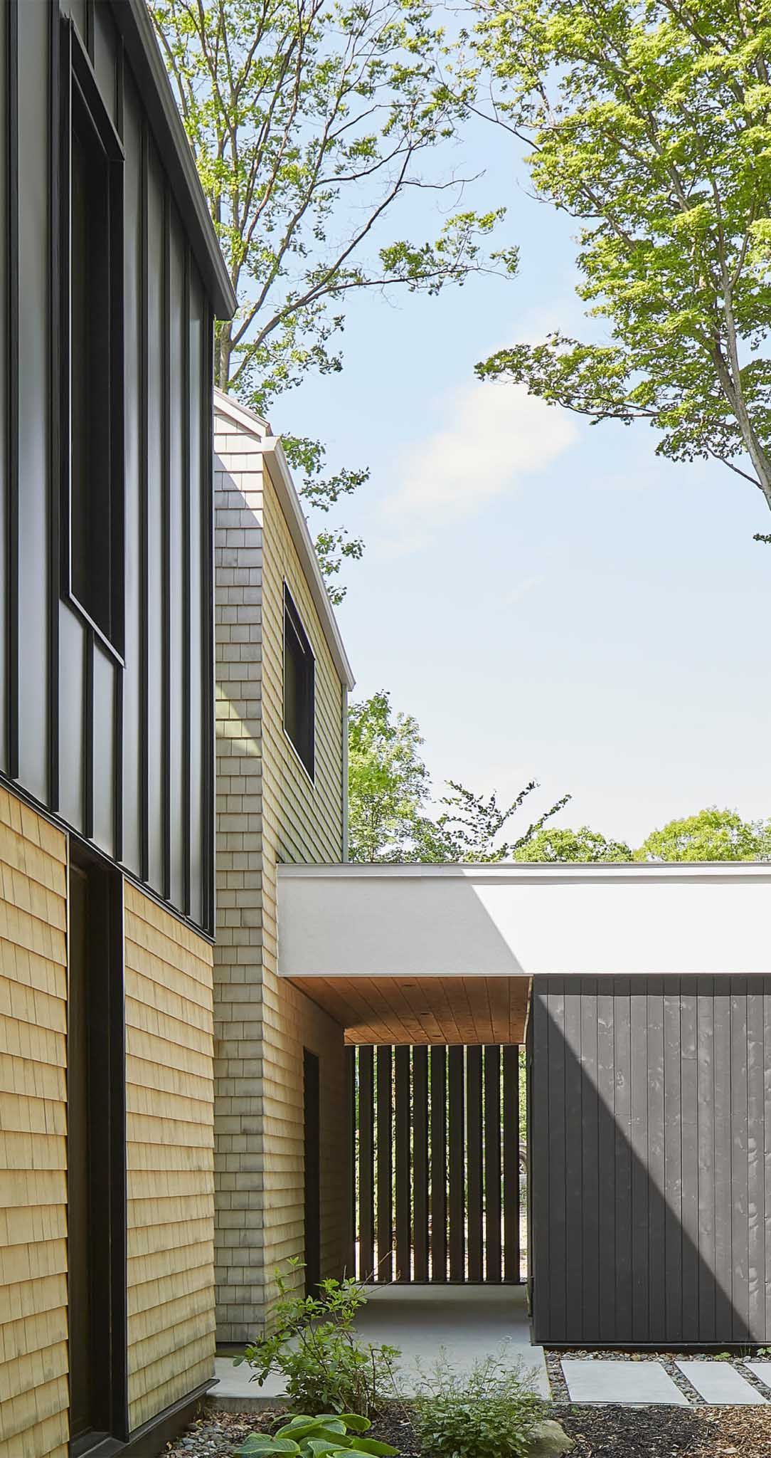 Maison moderne avec bardage fonc&eacute; et bardeaux de bois, all&eacute;e pi&eacute;tonne entre les b&acirc;timents.