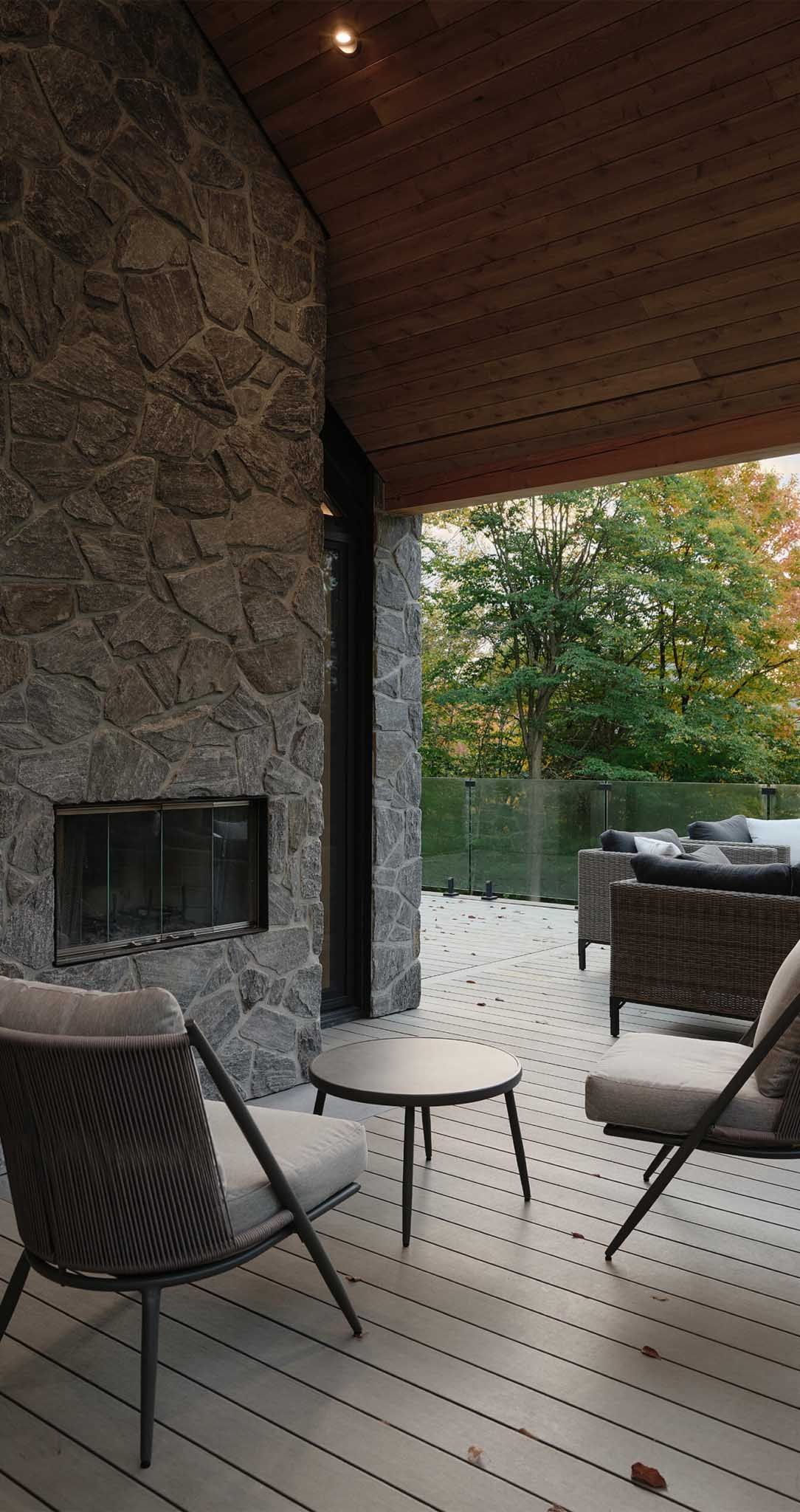 Espace ext&eacute;rieur am&eacute;nag&eacute; avec chemin&eacute;e en pierre, terrasse en bois et vue sur les arbres.