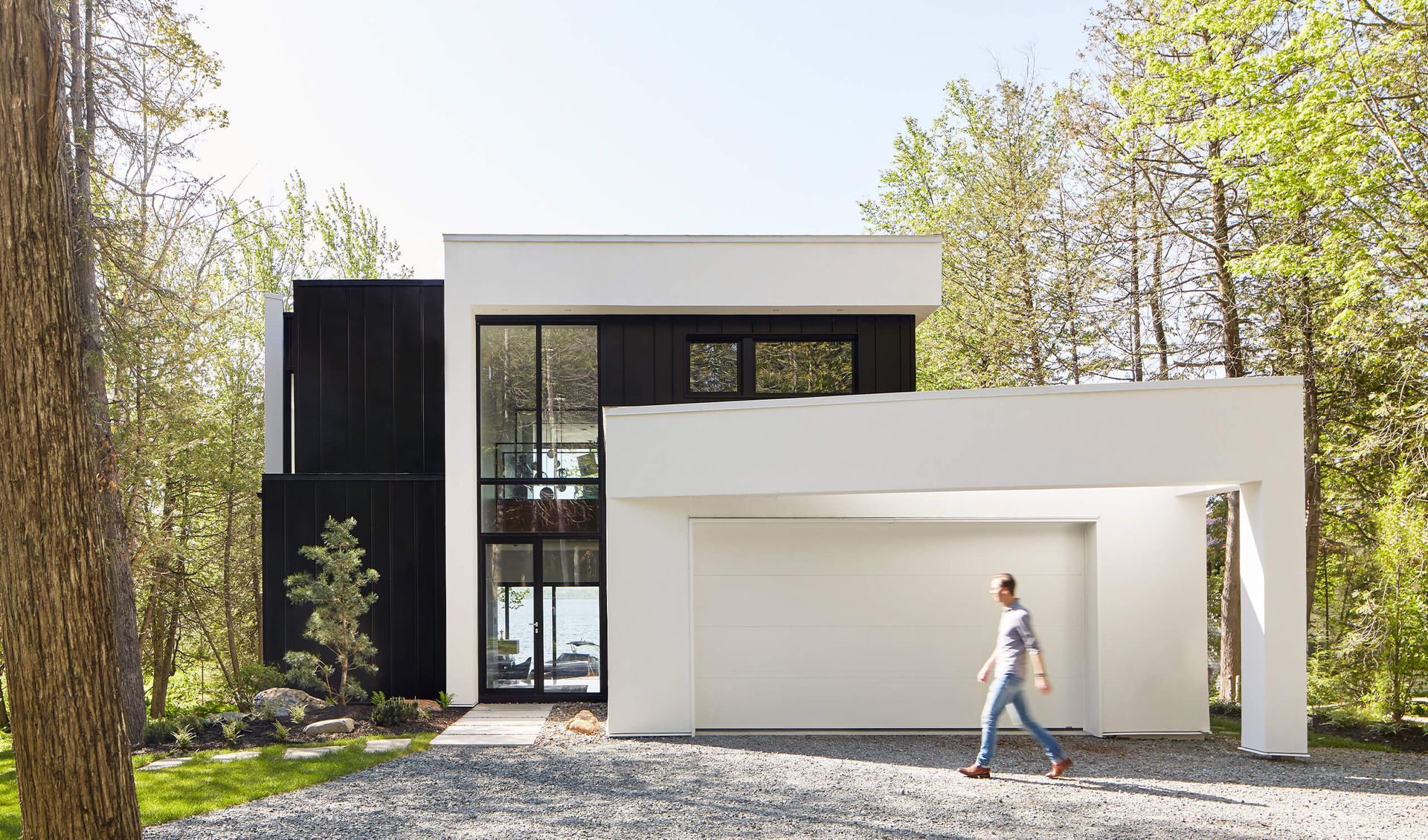 Maison moderne en noir et blanc avec une femme passant devant un abri de voiture par une journée ensoleillée.