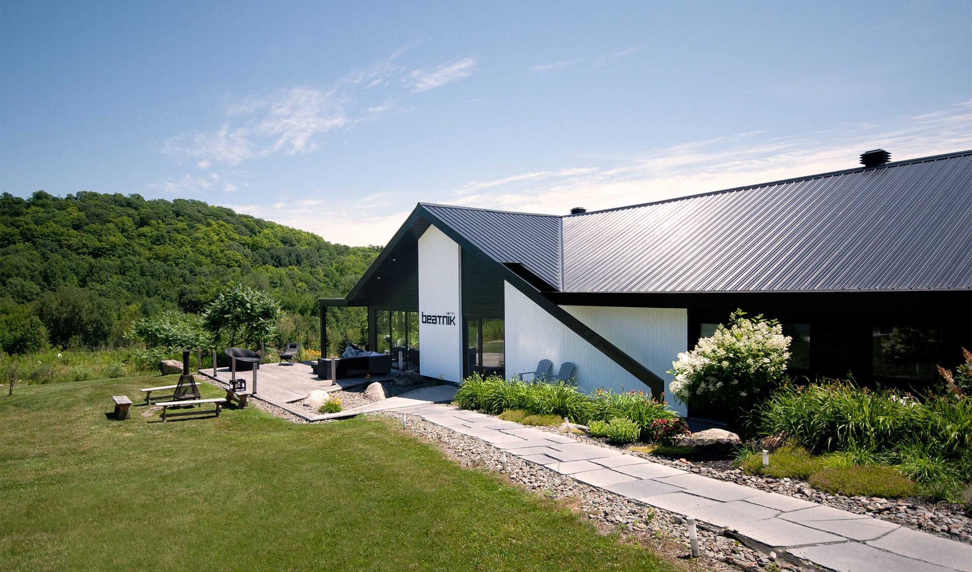 Maison moderne au toit noir et aux murs blancs, perchée sur une colline herbeuse ; arbres en arrière-plan, ciel bleu.