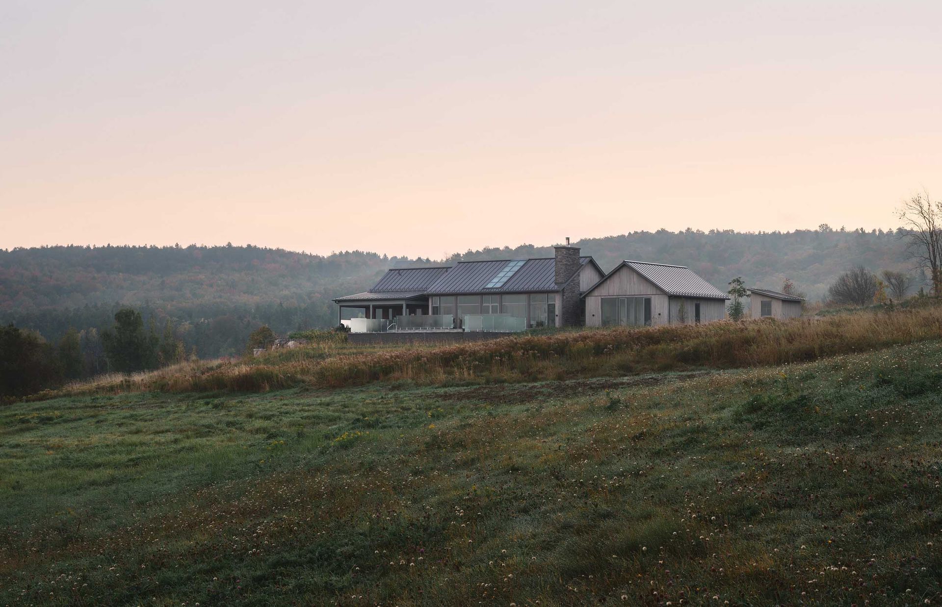 Un b&acirc;timent de plain-pied se dresse sur une colline herbeuse, avec une for&ecirc;t en arri&egrave;re-plan ; un ciel aux teintes douces au-dessus.
