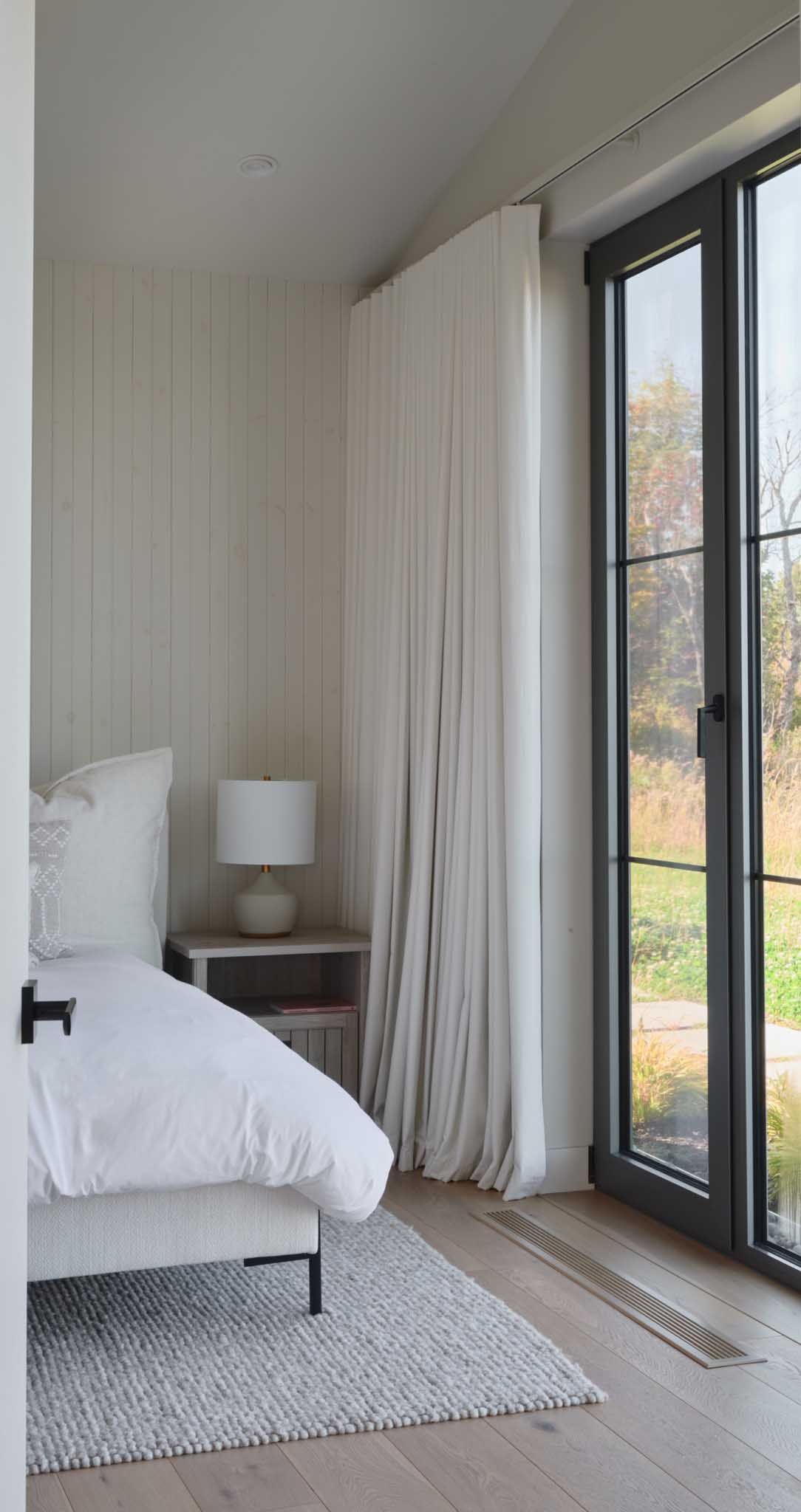 Chambre avec murs blancs, lit et voilages pr&egrave;s d'une grande fen&ecirc;tre.