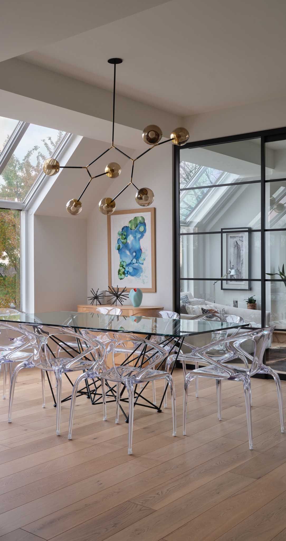 Salle &agrave; manger avec table en verre, chaises transparentes, lustre moderne, &oelig;uvres d'art et puits de lumi&egrave;re.