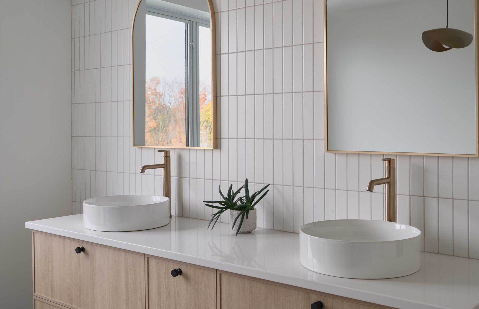 Salle de bains avec murs carrel&eacute;s blancs, deux lavabos sur un meuble-lavabo en bois et miroirs &agrave; cadre dor&eacute;.