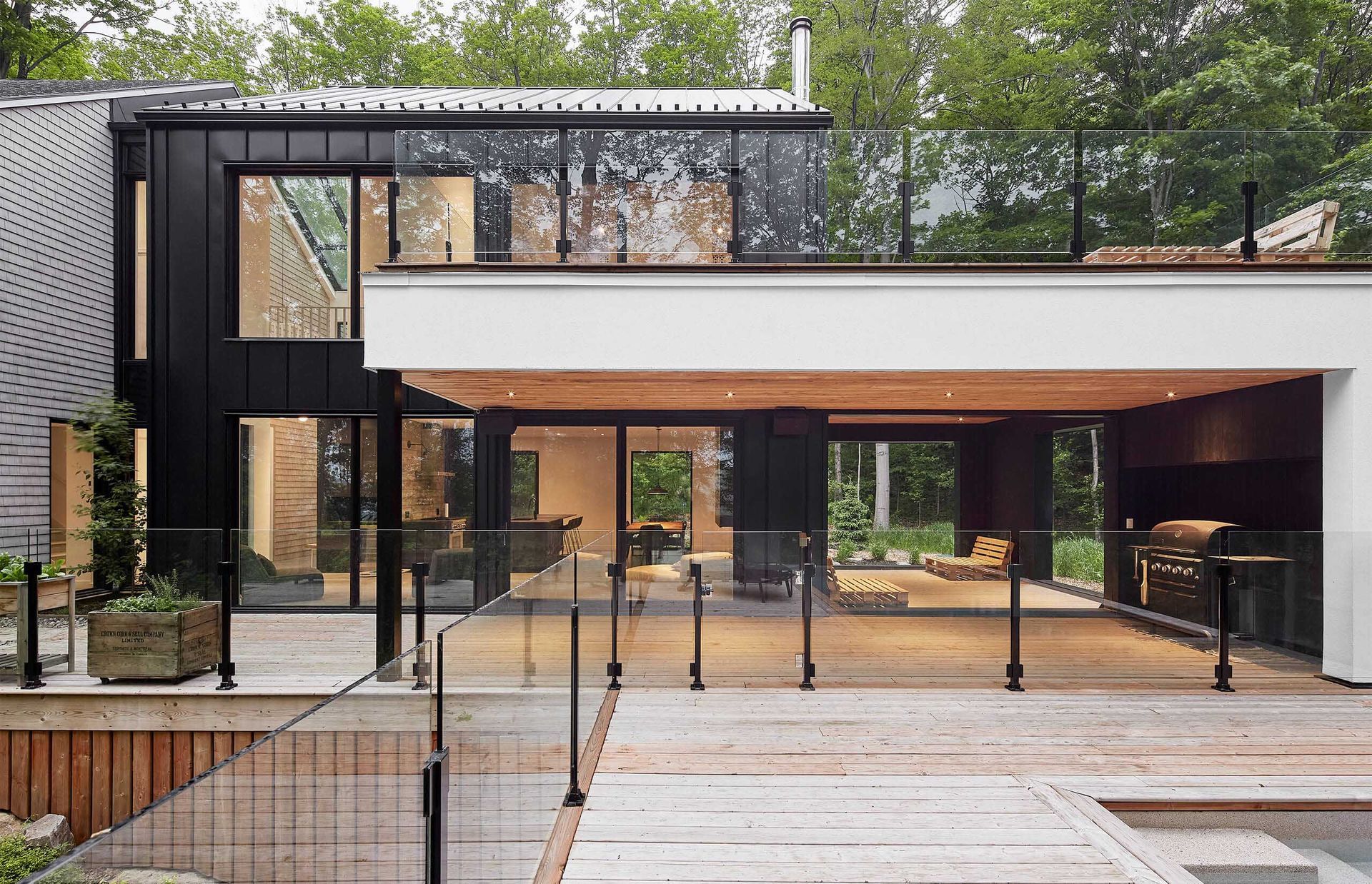 Modern house with black exterior, glass walls, wooden deck, and a grill area.