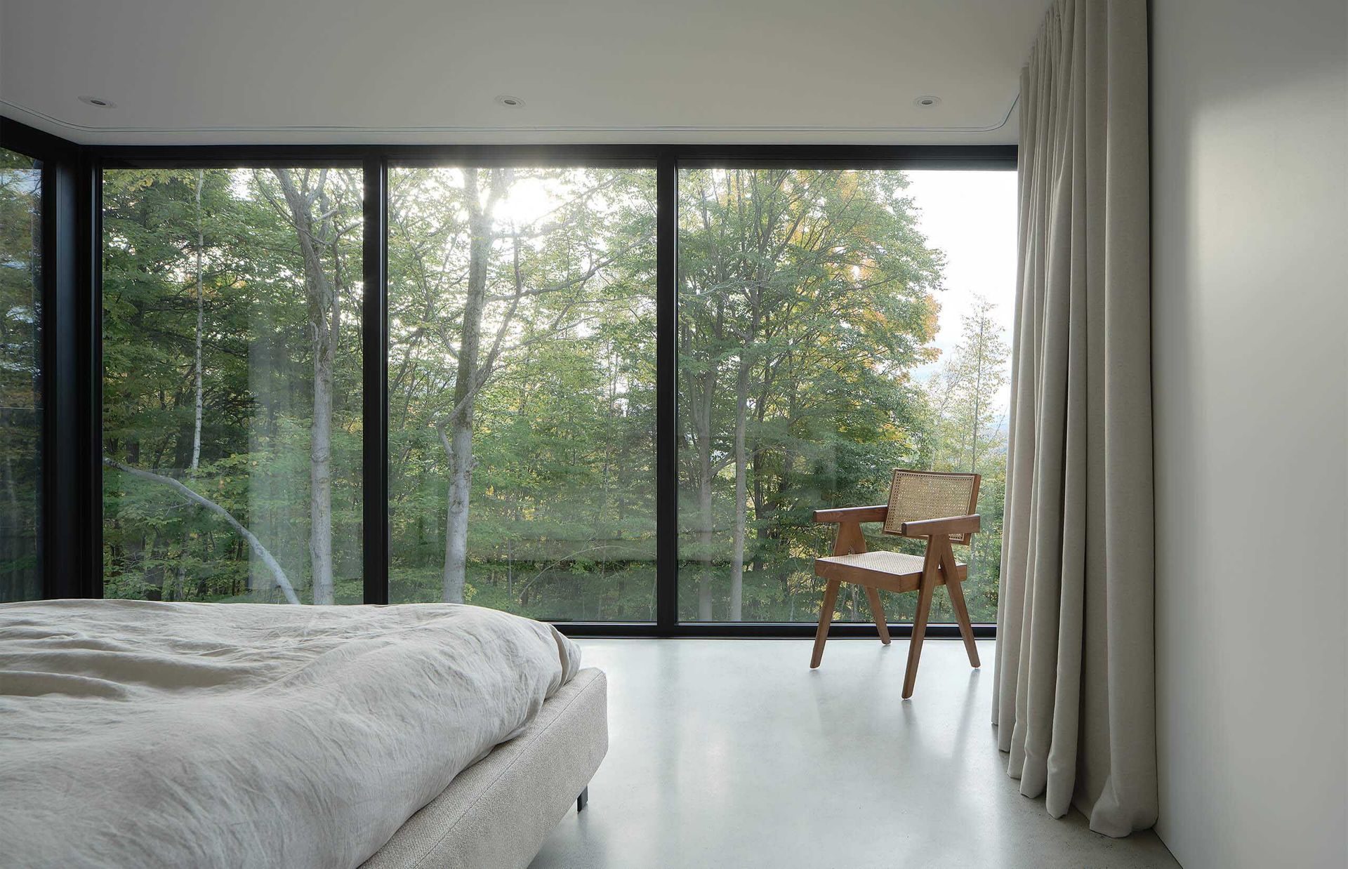 Chambre avec une grande fen&ecirc;tre donnant sur les arbres ; une chaise est plac&eacute;e devant la fen&ecirc;tre, le lit au premier plan.