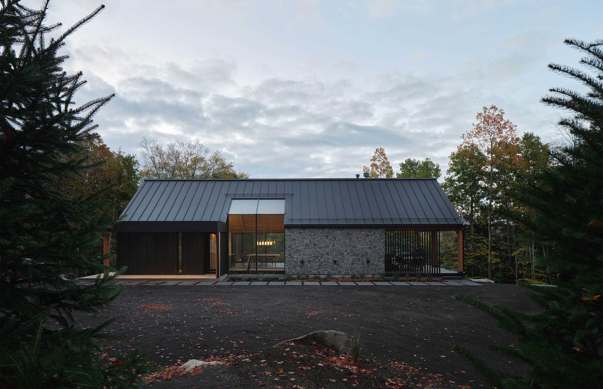 Cabane moderne au toit noir et &agrave; la fa&ccedil;ade en pierre, au milieu des arbres sous un ciel nuageux.