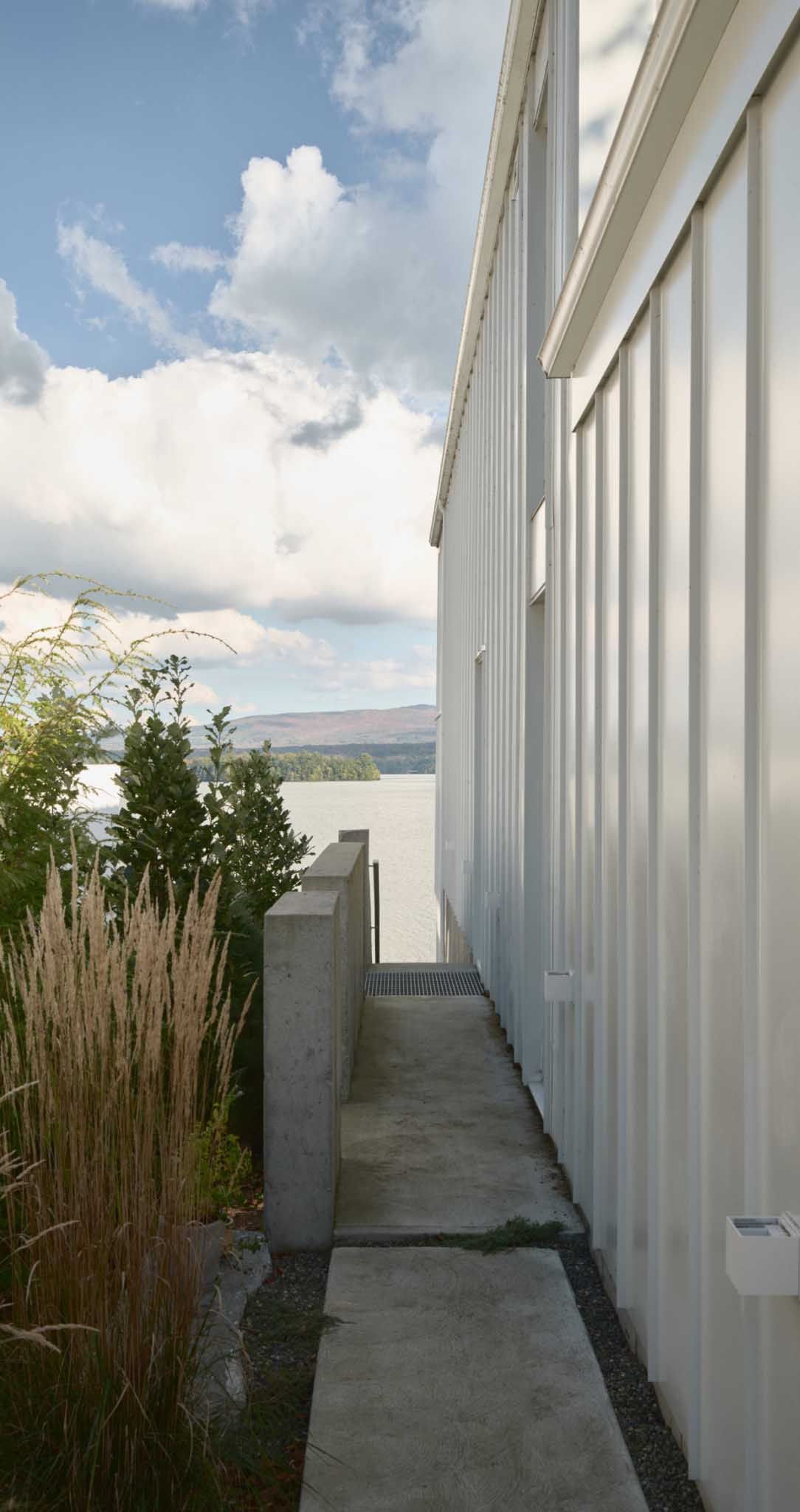 B&acirc;timent blanc &agrave; bardage vertical, jouxtant une all&eacute;e pi&eacute;tonne en b&eacute;ton, donnant sur un plan d'eau sous un ciel nuageux.