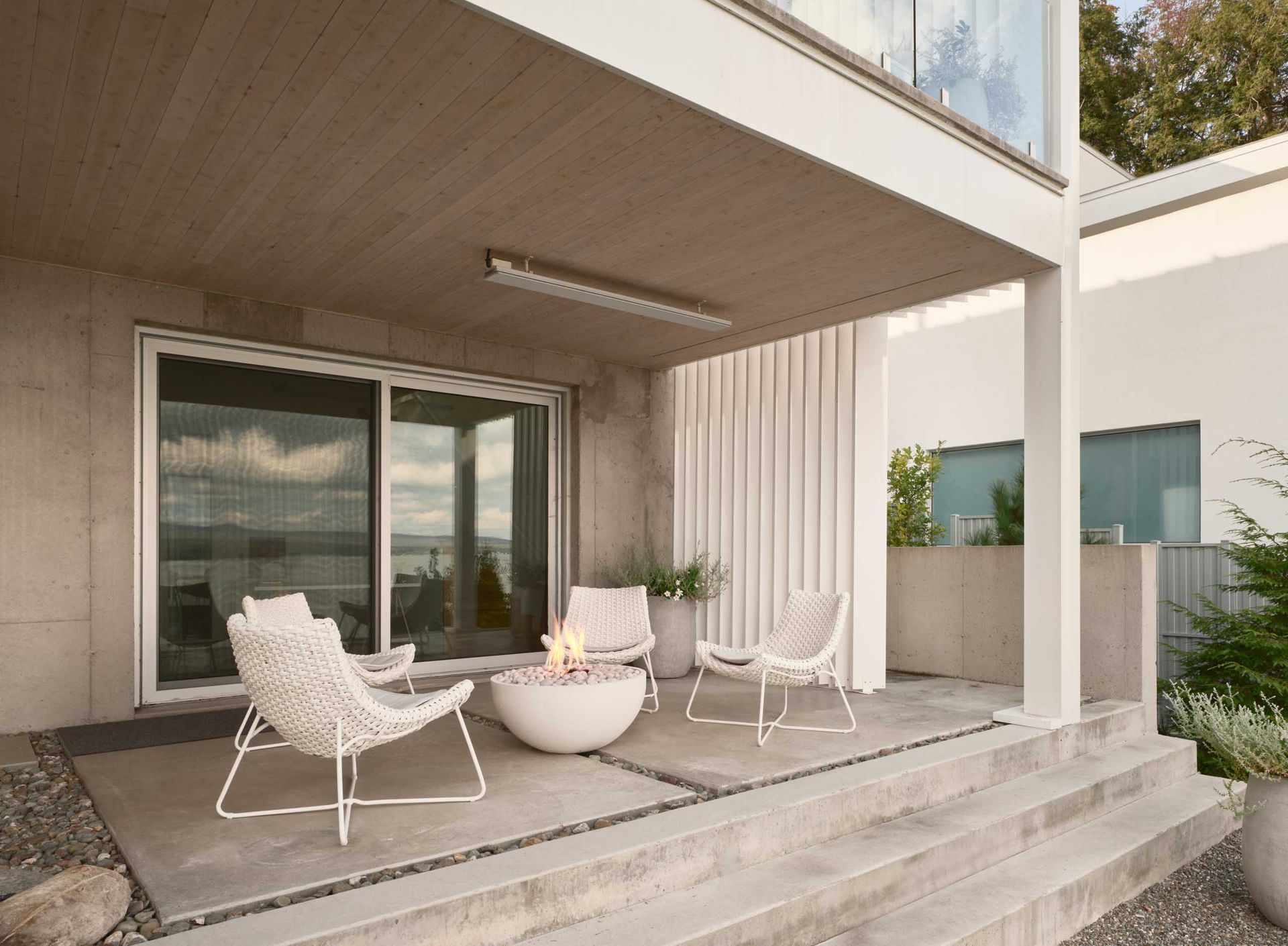 Terrasse avec mobilier blanc, foyer ext&eacute;rieur et portes coulissantes donnant sur un lac.