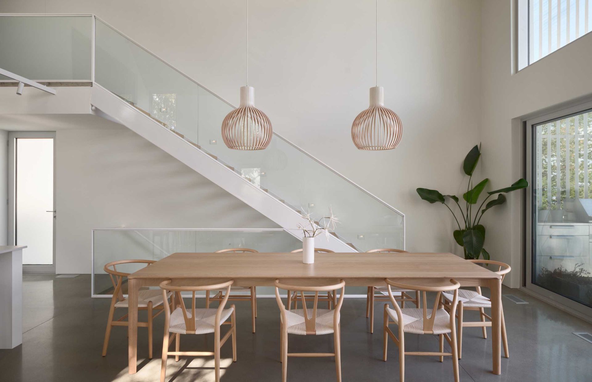 Light-filled dining room with wood table, chairs, and two pendant lights. A white staircase and large window are visible.