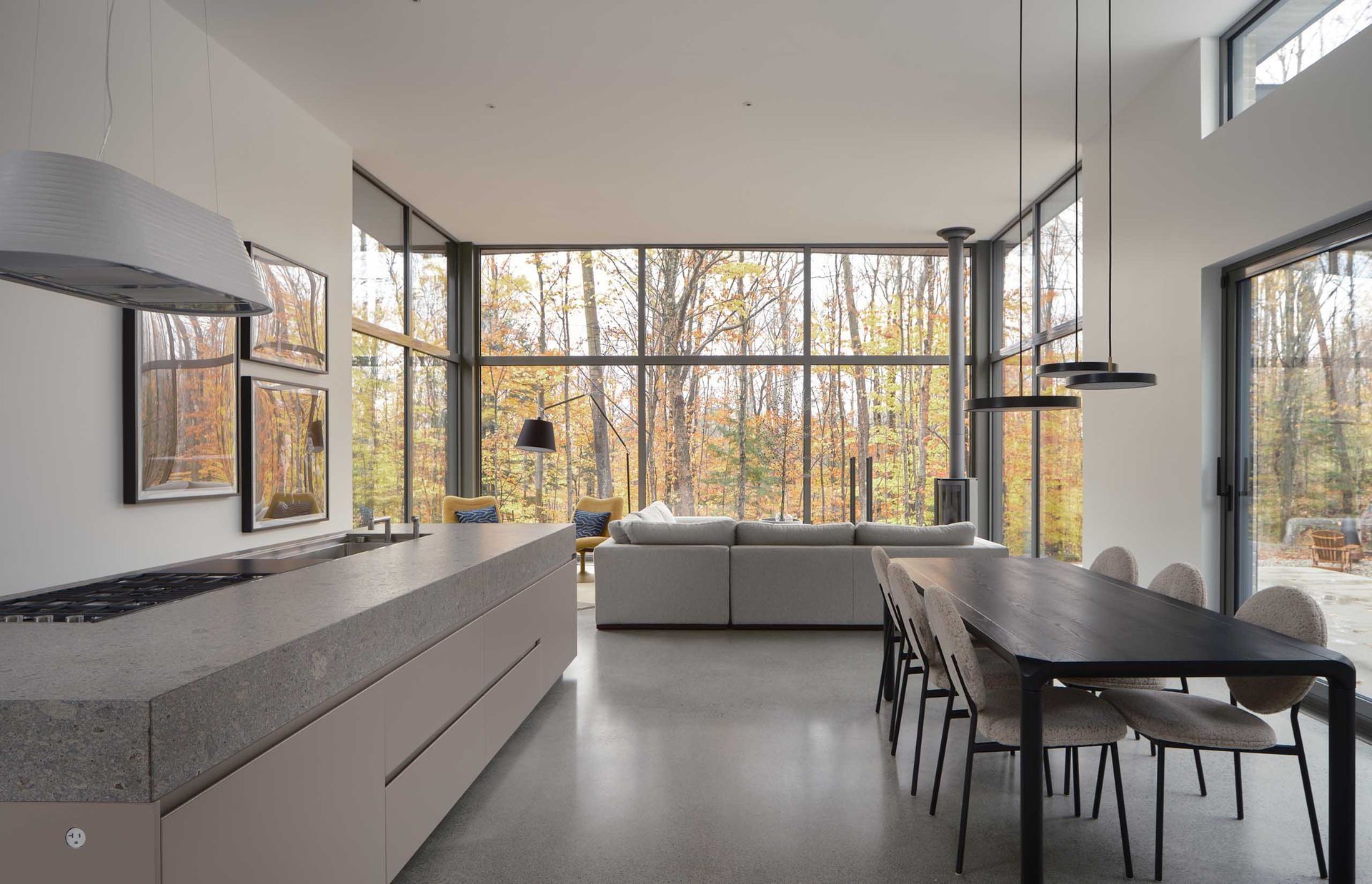 Cuisine et salle &agrave; manger modernes et ouvertes, baign&eacute;es de lumi&egrave;re gr&acirc;ce &agrave; de grandes fen&ecirc;tres donnant sur la for&ecirc;t. Murs et sols gris clair, design minimaliste.