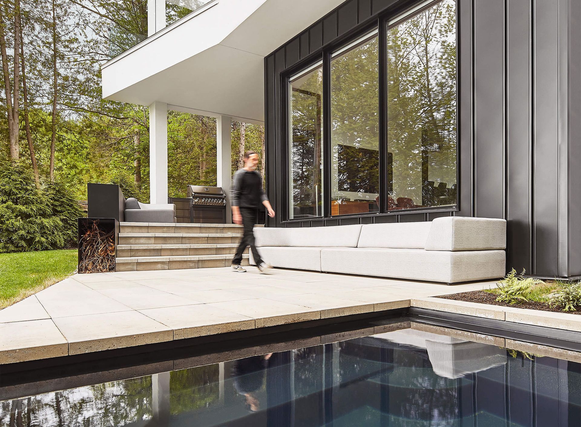 Une personne se prom&egrave;ne sur une terrasse &agrave; c&ocirc;t&eacute; d'une piscine et d'une maison moderne avec de grandes fen&ecirc;tres.