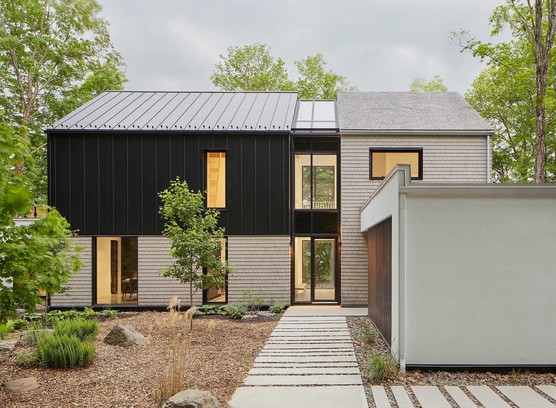 Maison moderne &agrave; deux &eacute;tages avec bardage noir et fa&ccedil;ade en pierre textur&eacute;e, entour&eacute;e d'arbres et d'un chemin en pierre.