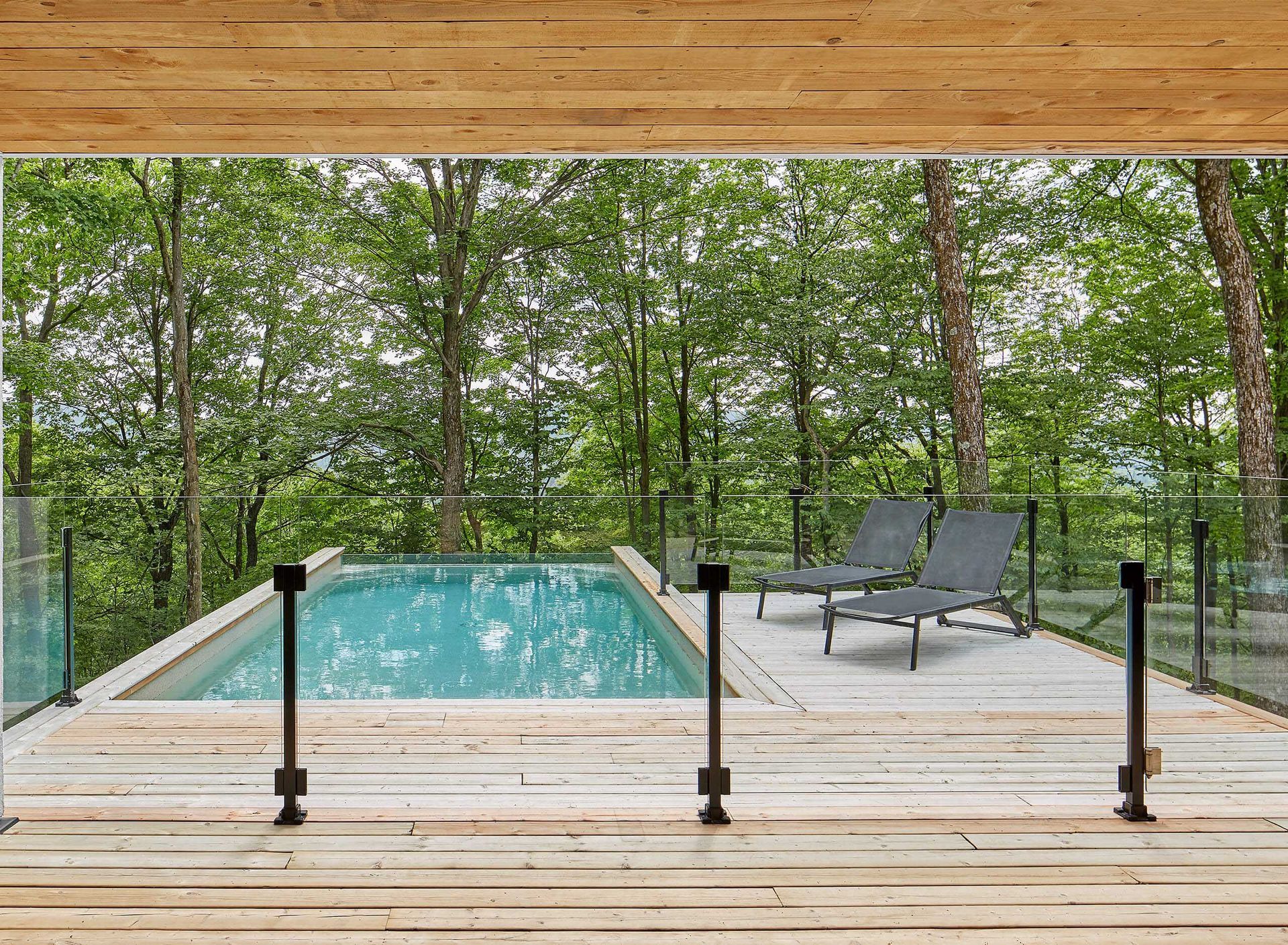 Vue depuis la piscine: piscine bleue, terrasse avec chaises longues, balustrade en verre, for&ecirc;t en arri&egrave;re-plan, plafond en bois.