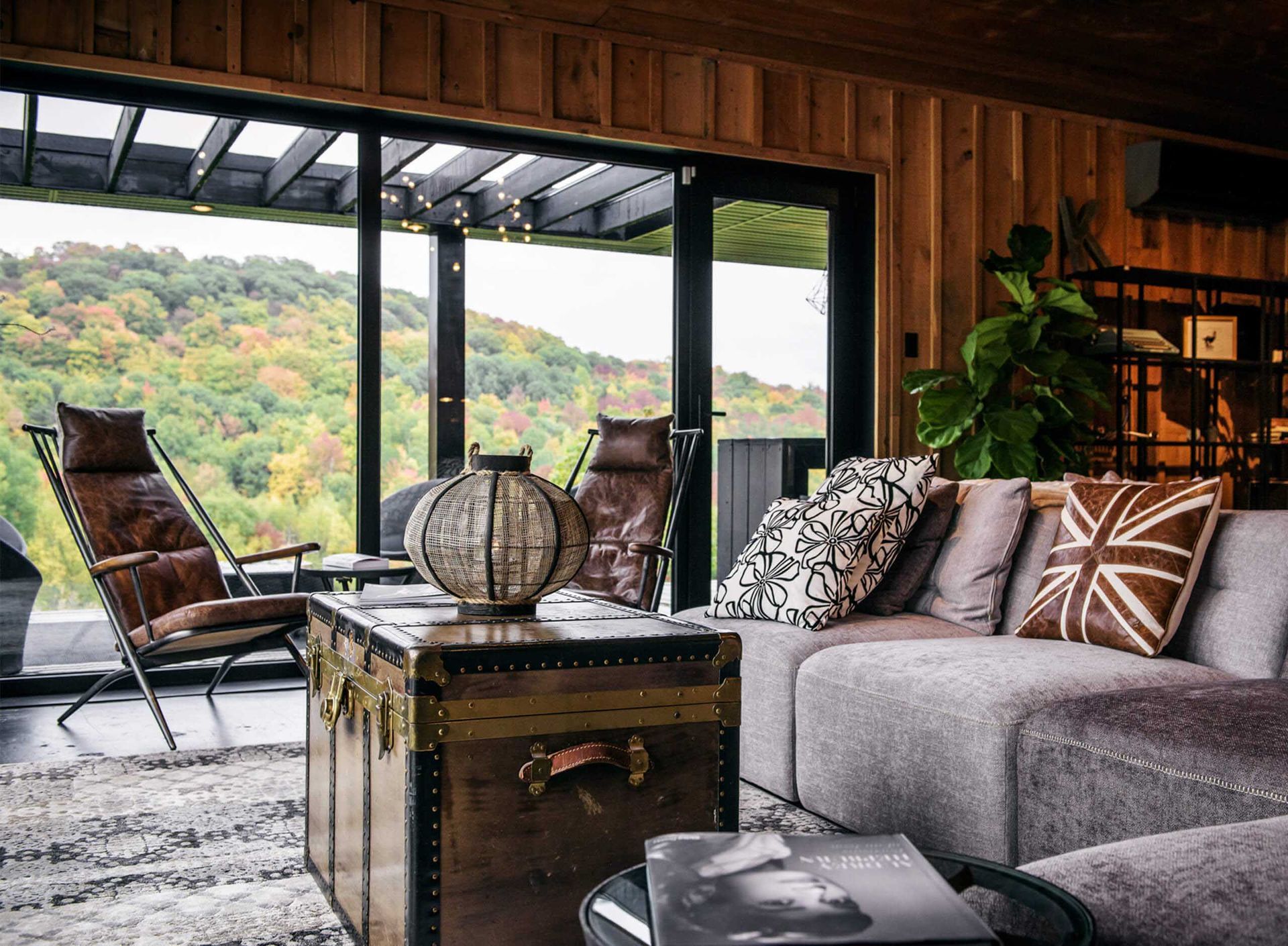 Salon confortable de chalet avec vue sur la for&ecirc;t. Fauteuils en cuir, coussins &agrave; motifs, table basse en forme de coffre et grandes portes coulissantes.