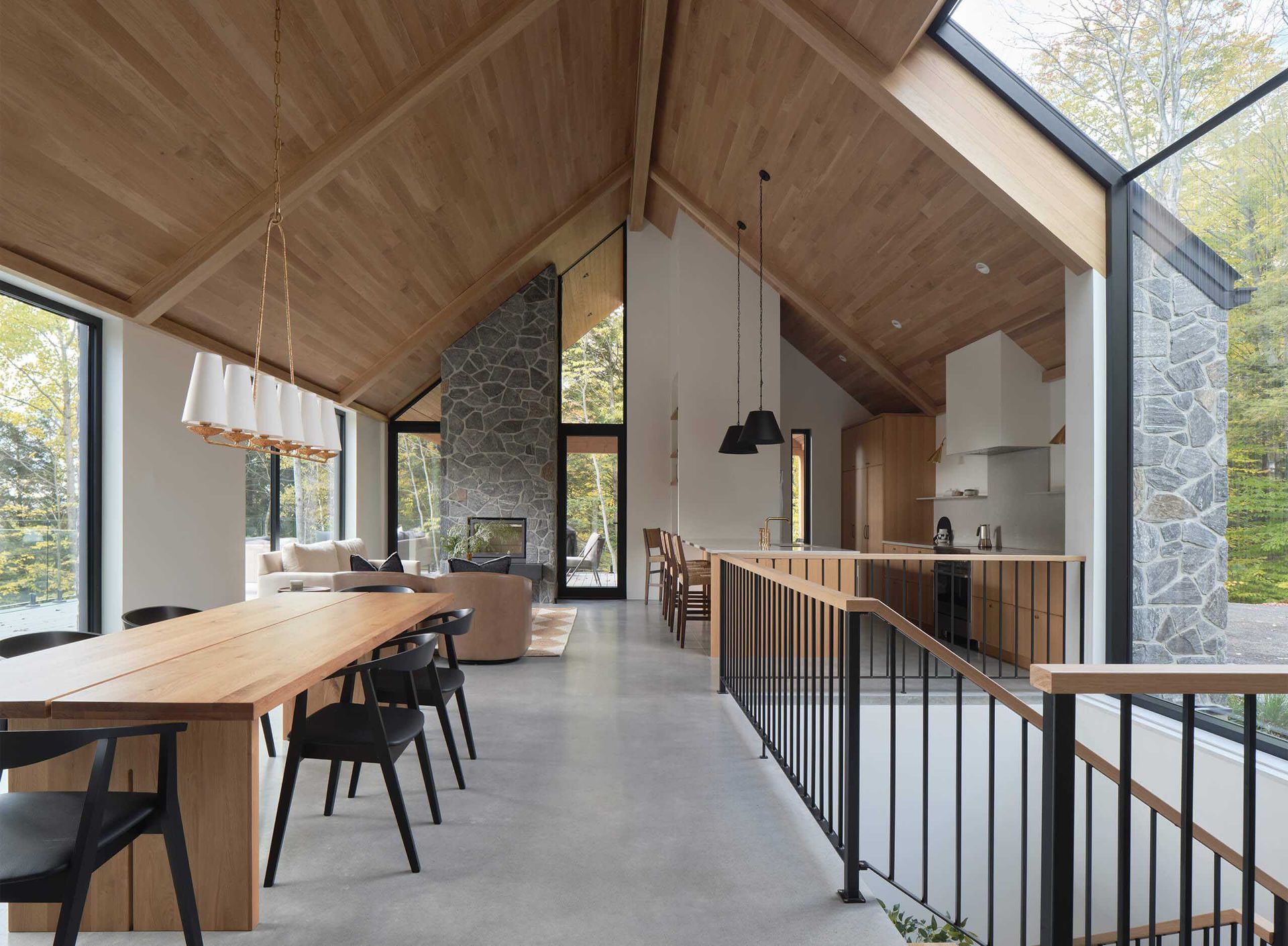Int&eacute;rieur d'une maison moderne avec plafond lambriss&eacute;, grandes fen&ecirc;tres, table &agrave; manger et chemin&eacute;e en pierre.