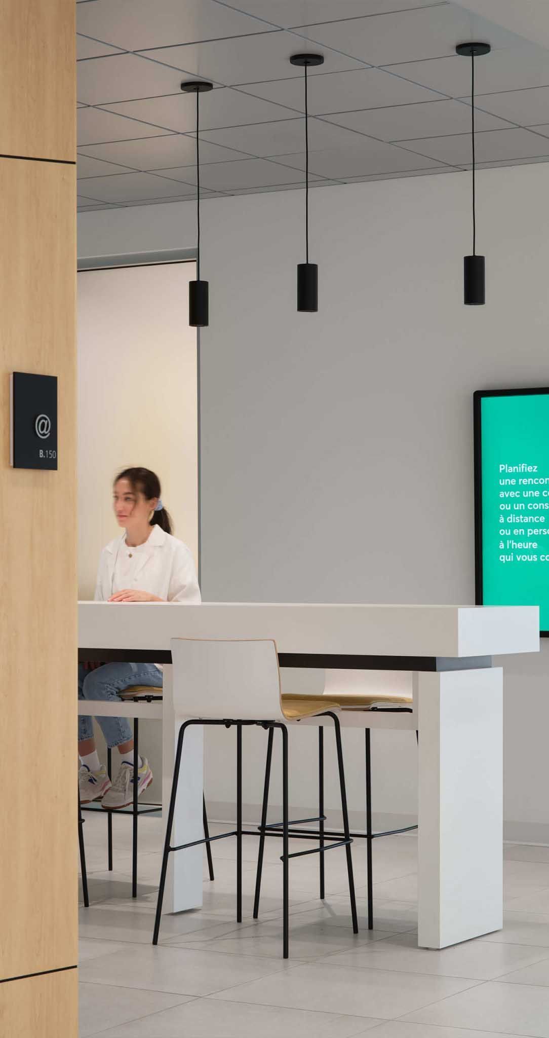 Une femme est assise &agrave; un comptoir blanc dans un bureau moderne, avec des tabourets. Un luminaire noir est suspendu au-dessus.