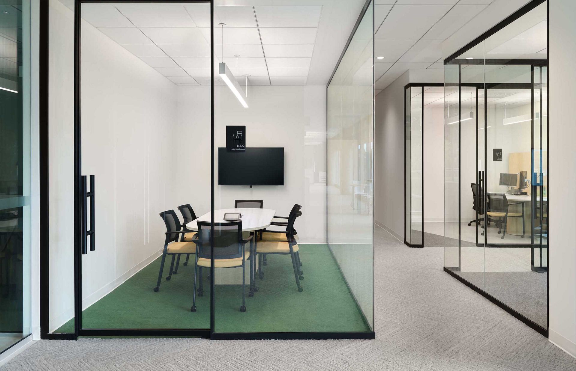 Espace de bureaux moderne avec salle de conf&eacute;rence et couloir vitr&eacute;s. Moquette verte, murs blancs, cadres noirs.