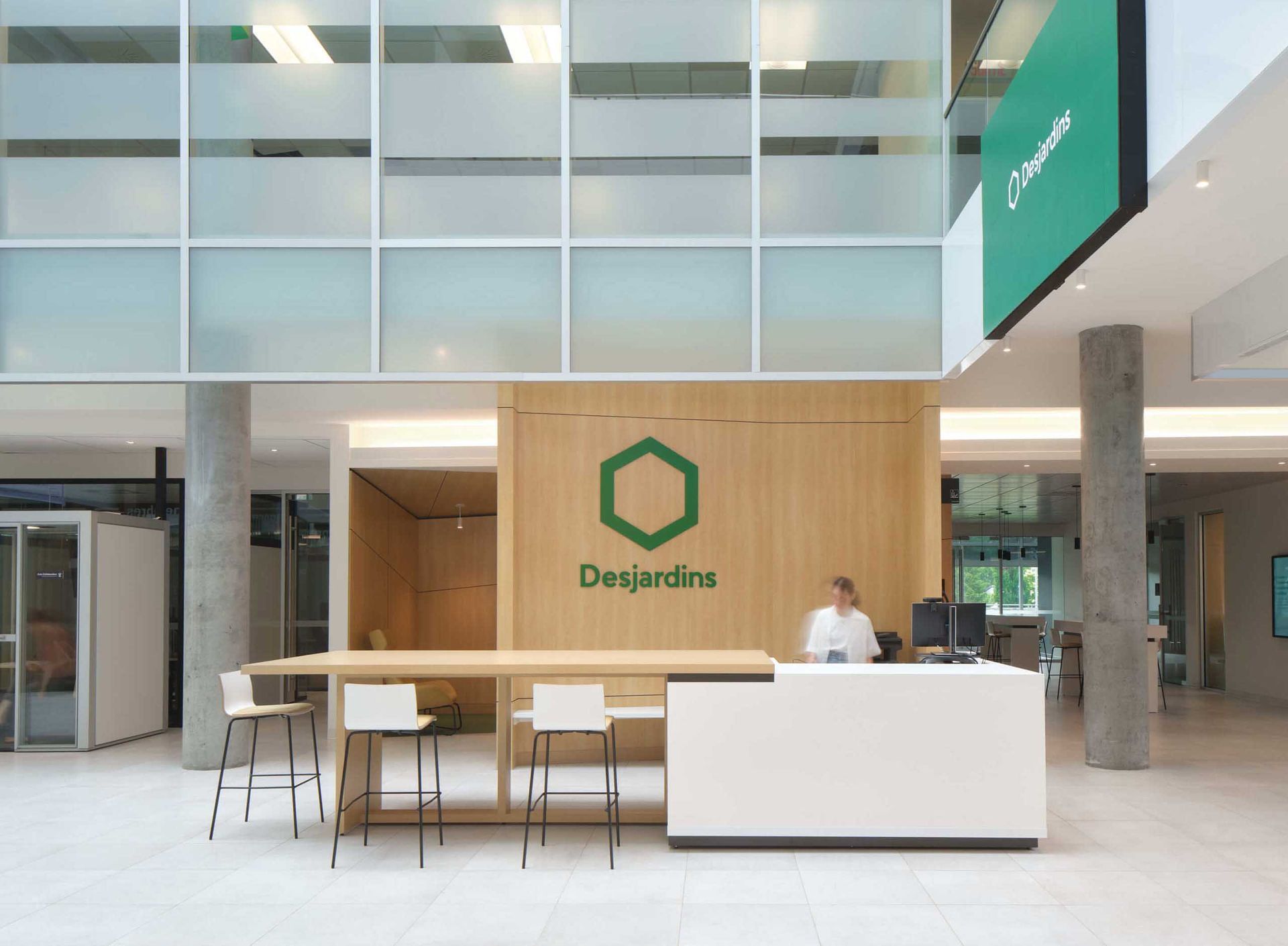 Espace d'accueil avec le logo Desjardins, des boiseries, un comptoir blanc et une personne assise derri&egrave;re.