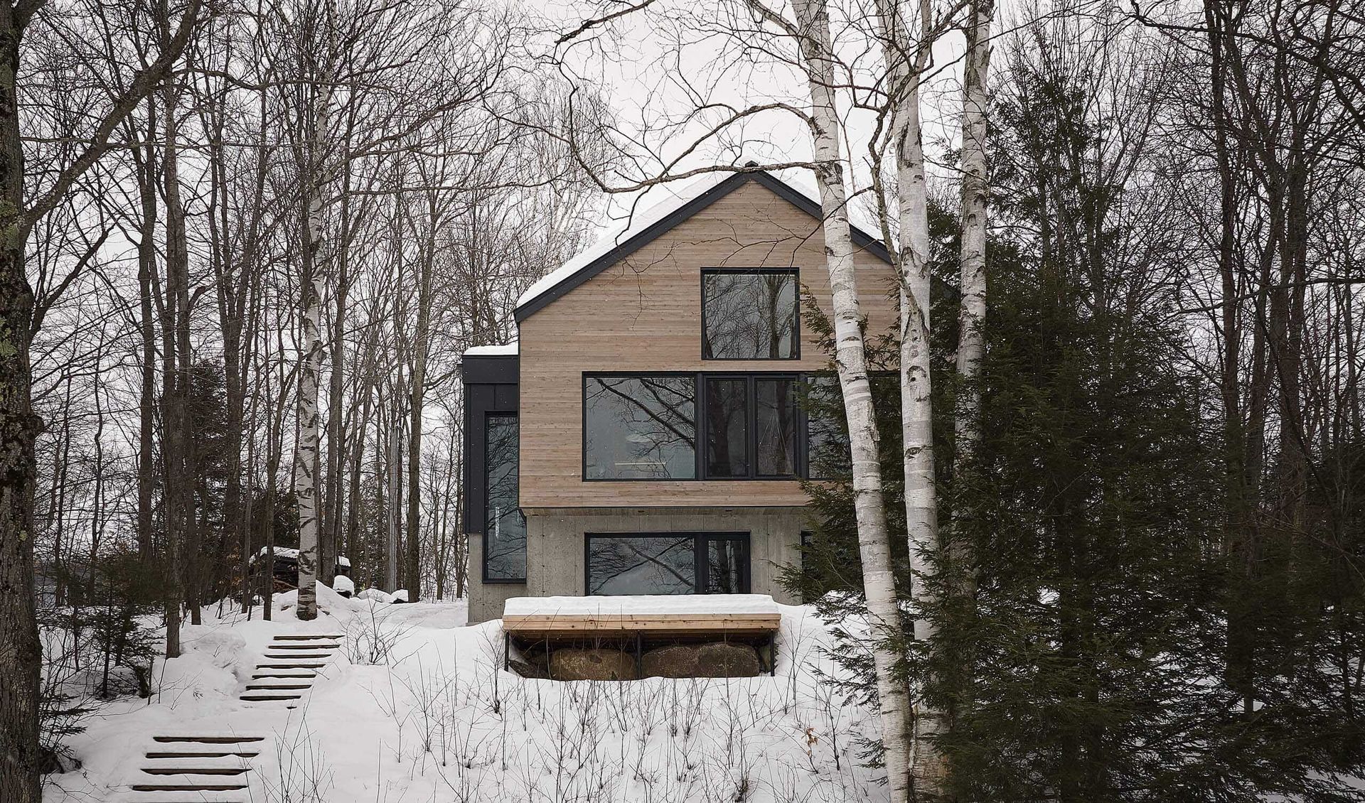Maison moderne en forêt enneigée, avec de grandes fenêtres et un bardage en bois. Un escalier mène à une terrasse.