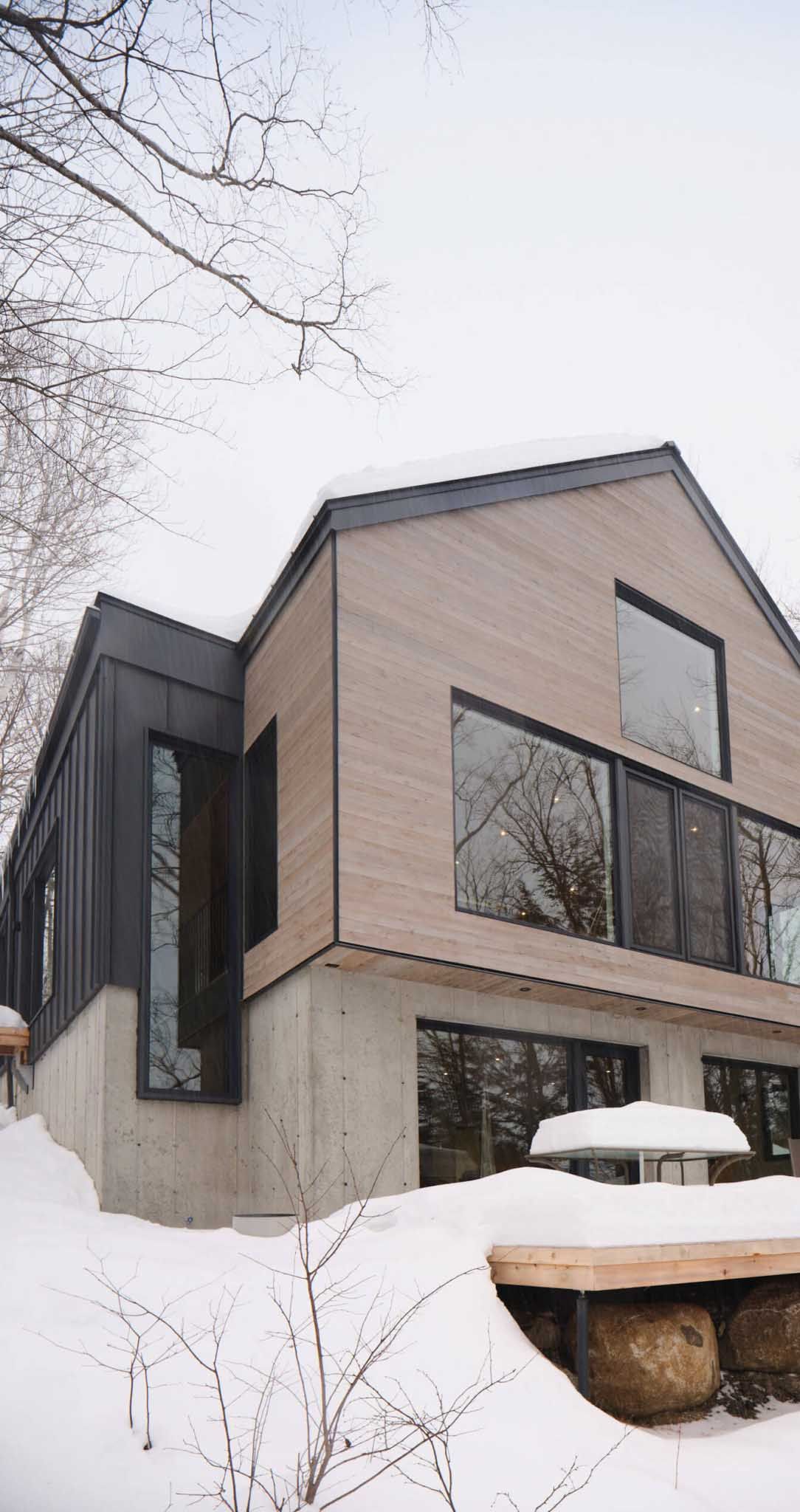 Maison moderne avec bardage en bois et grandes fen&ecirc;tres, entour&eacute;e de neige et d'arbres.
