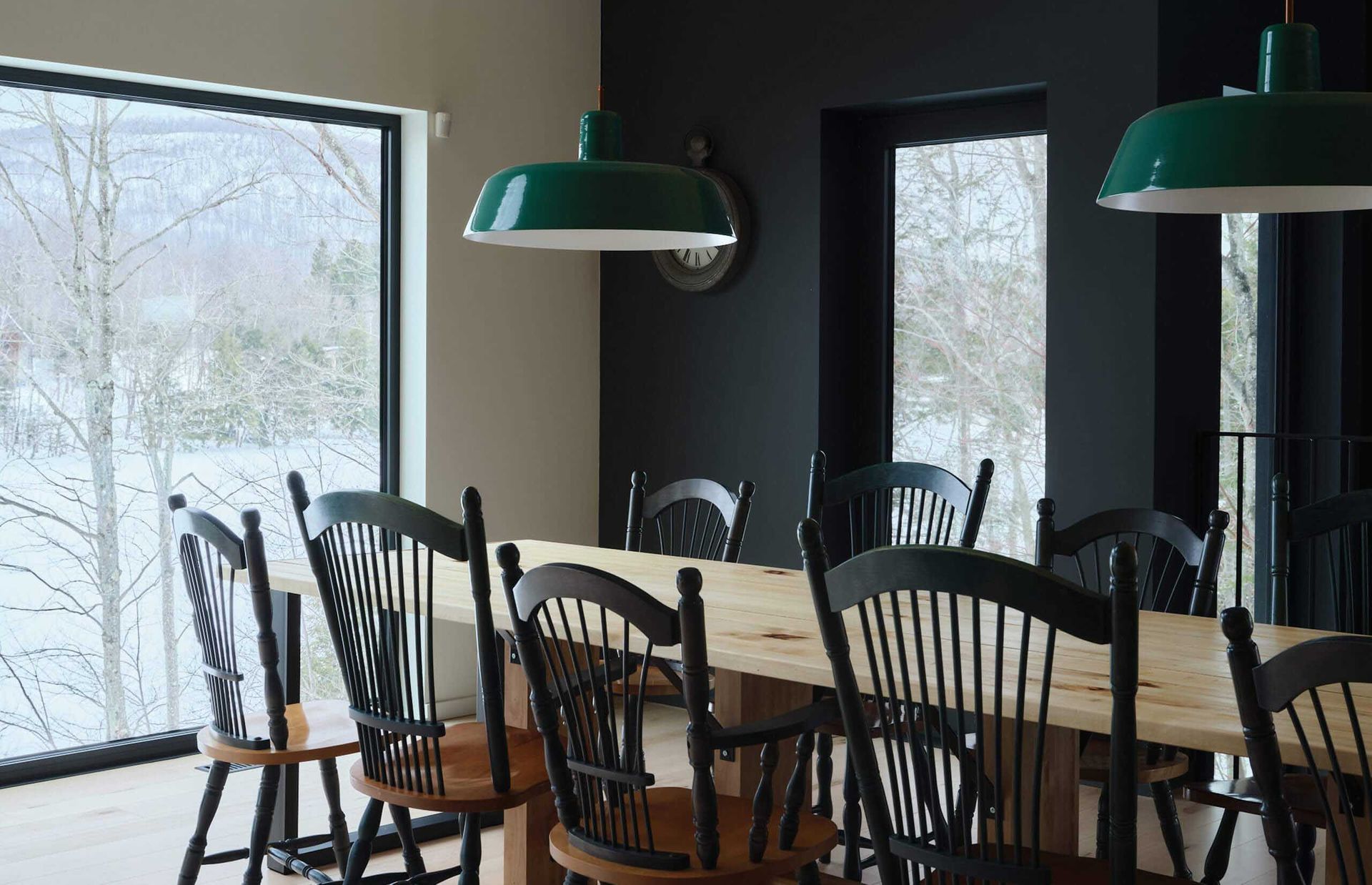 Salle &agrave; manger avec grande table en bois, chaises noires, suspensions vertes et vue sur la neige par la fen&ecirc;tre.
