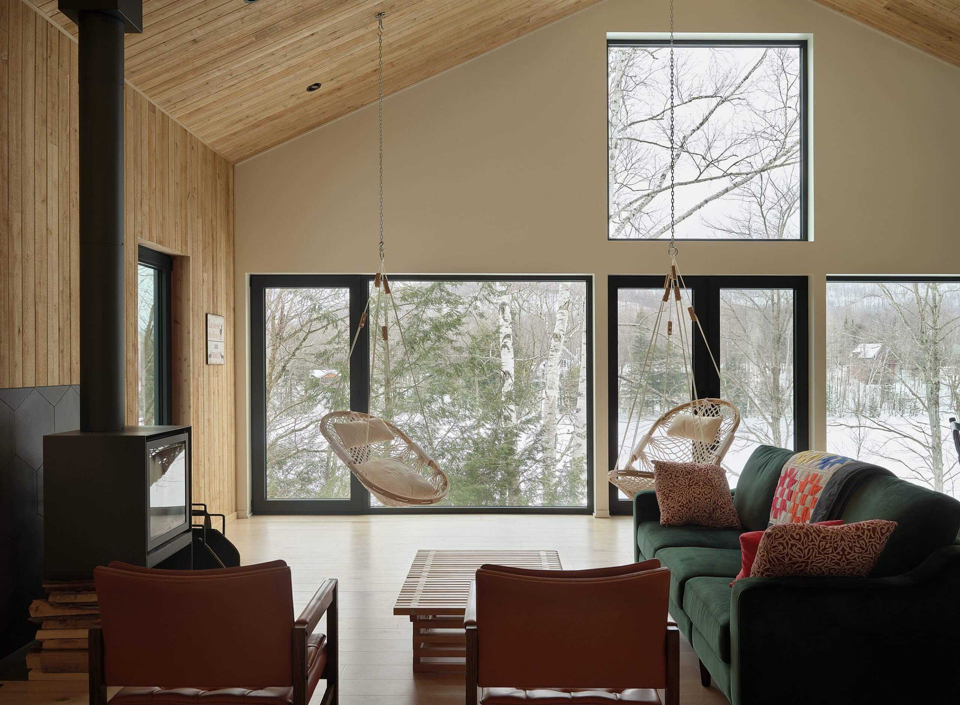 Salon moderne avec baies vitr&eacute;es donnant sur une for&ecirc;t enneig&eacute;e. Comprend une chemin&eacute;e, un canap&eacute; vert et des fauteuils suspendus.