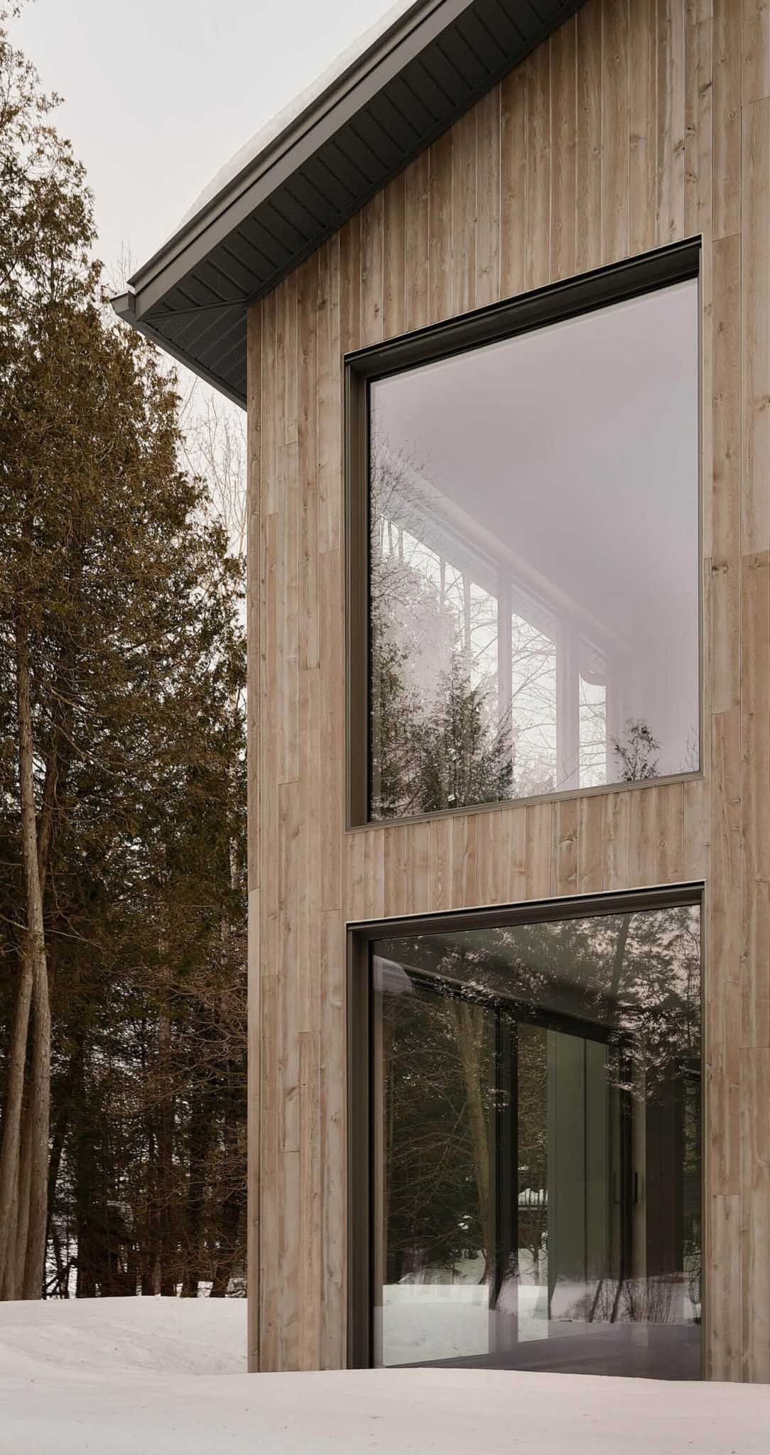 Immeuble imposant, rev&ecirc;tu de bois, avec de grandes fen&ecirc;tres refl&eacute;tant des arbres enneig&eacute;s, se d&eacute;tachant sur un paysage hivernal.